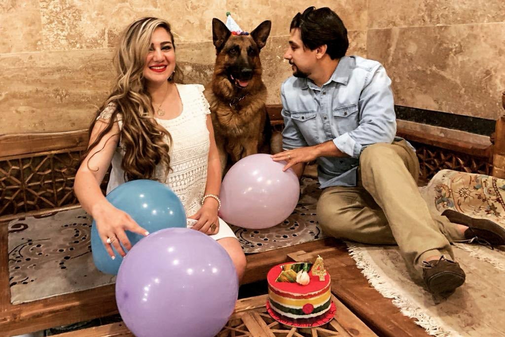Mahdis sits with her husband and a German shepherd wearing a party hat. It is a scene of celebration: All are smiling with balloons around them and a cake on the table in front of them.