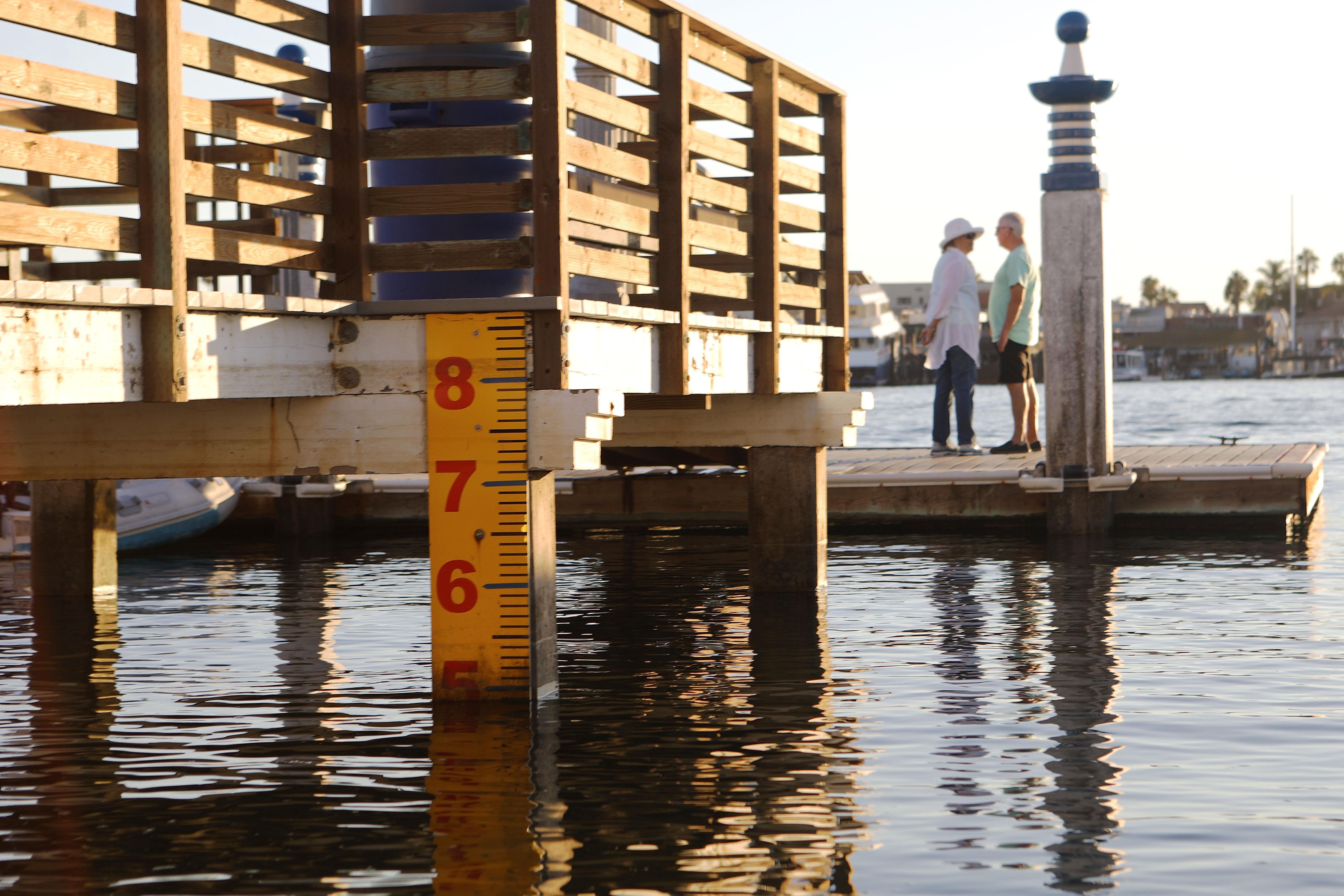 At a dock, water hits a water level marker at about 5 feet. Two people stand on the dock in the background.