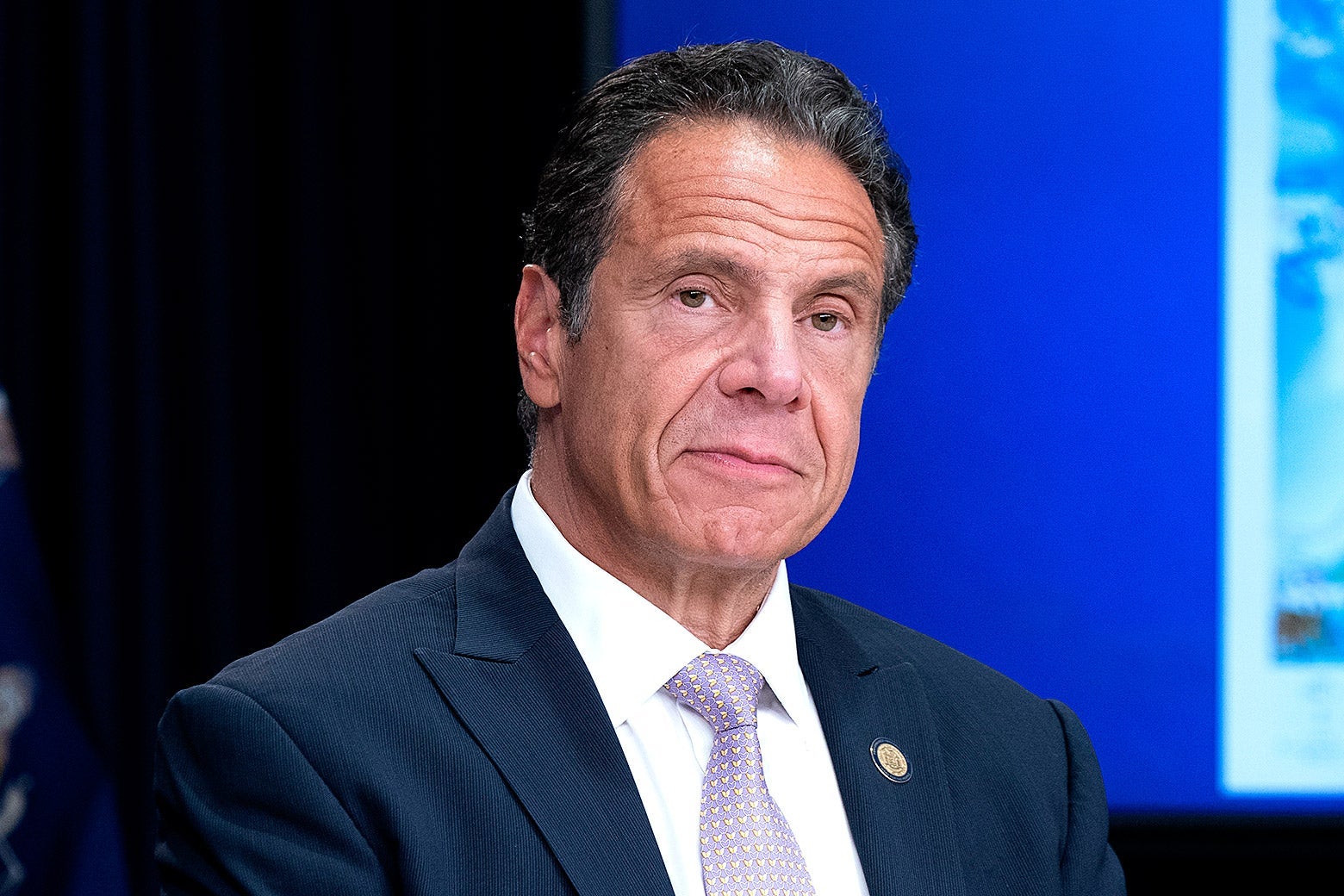 New York State Governor Andrew Cuomo sitting at a table with a serious expression while holding a media briefing.