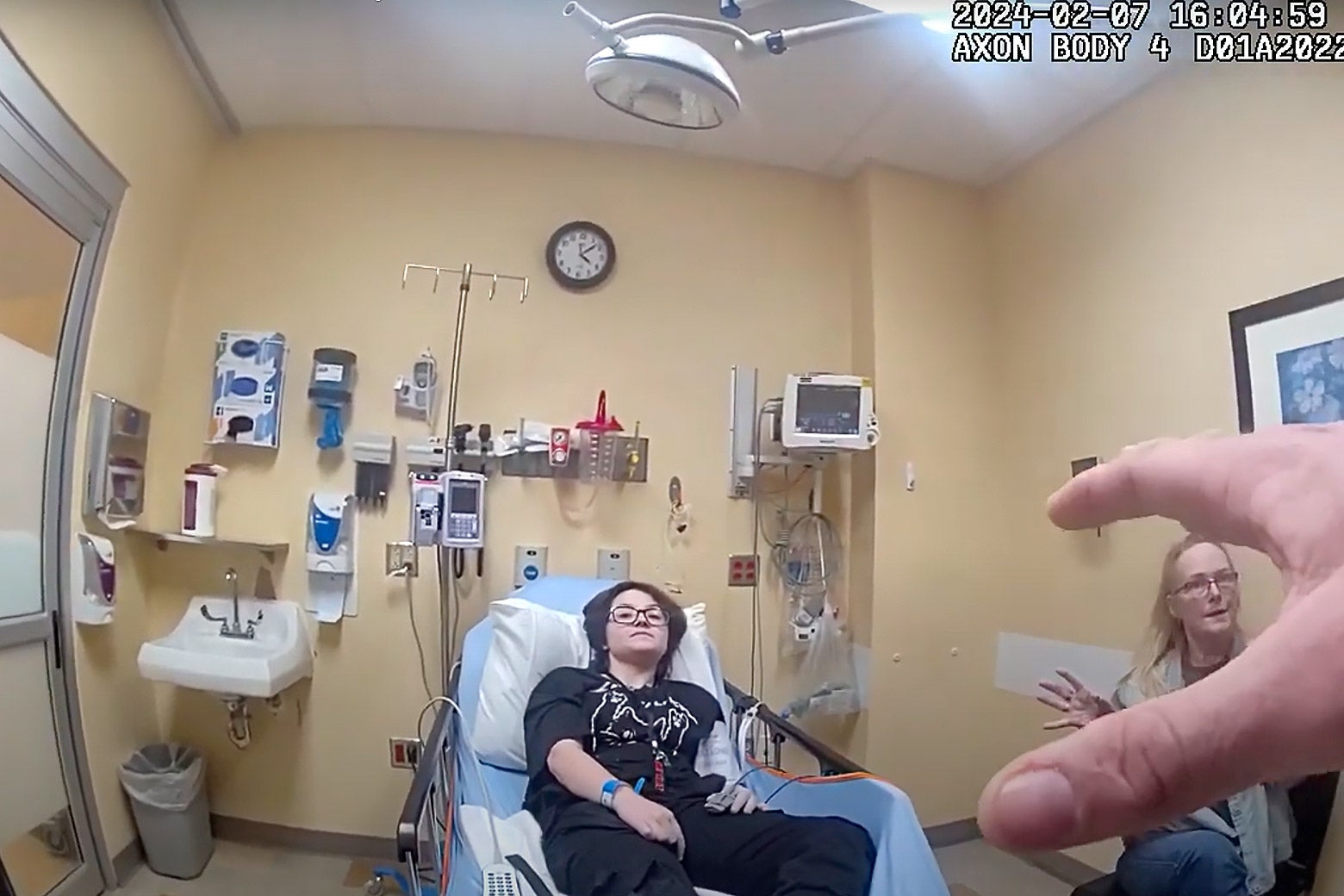 Nex Benedict is seen through a police body camera sitting back on a hospital bed. Nex is wearing all black and a woman sits nearby speaking to the officer, whose hand is in the frame. 