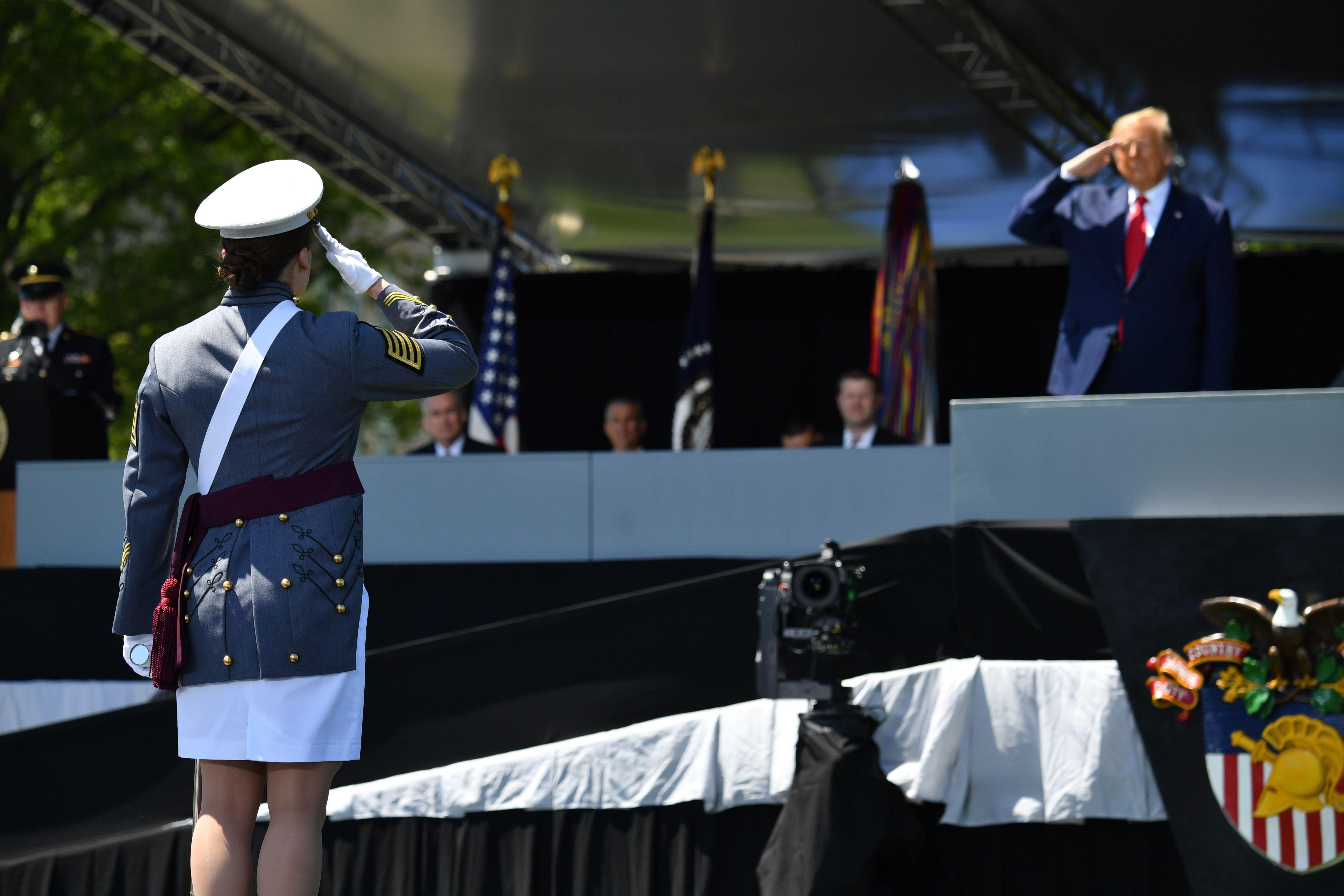 A female cadet with her back to the camera salutes Trump, who is standing onstage.