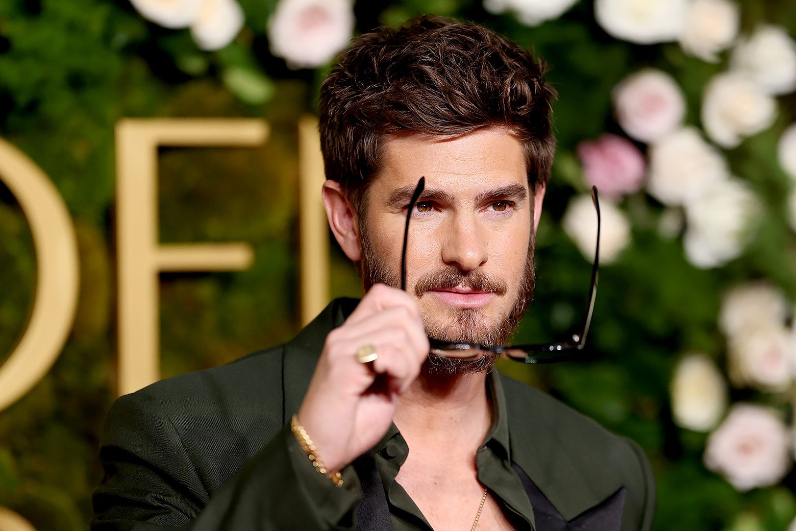 A fortysomething white man with a beard wears a button-down dark green shirt with a chain and puts sunglasses on, looking cool.