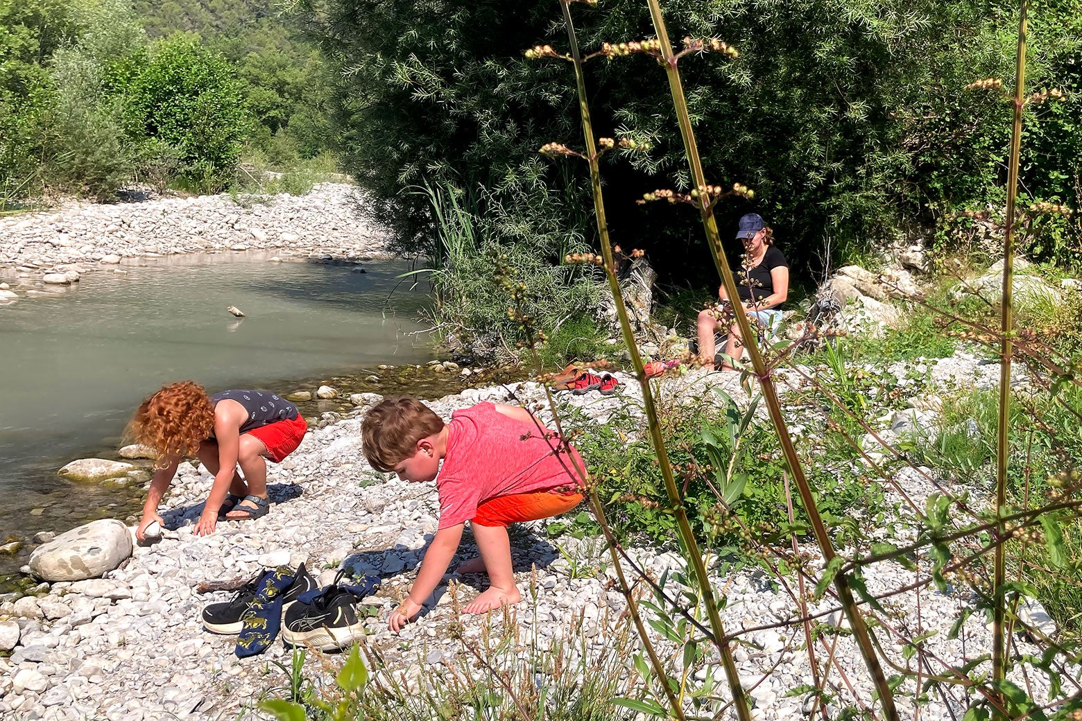 Two children play near a shallow river while a woman sits under a shade tree.