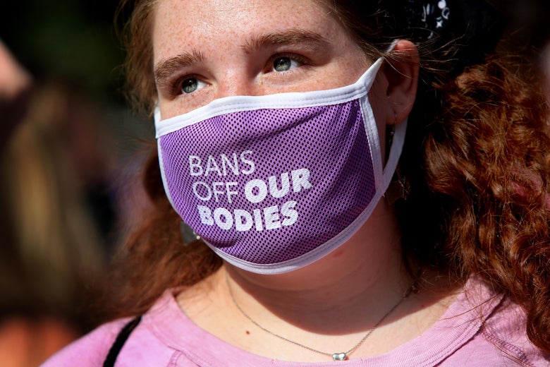 A person with red curly hair wears a mask that says "Bans Off Our Bodies."