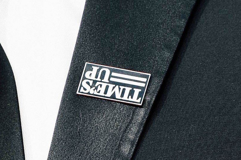An upside down Time’s Up pin on the lapel of a suit jacket.