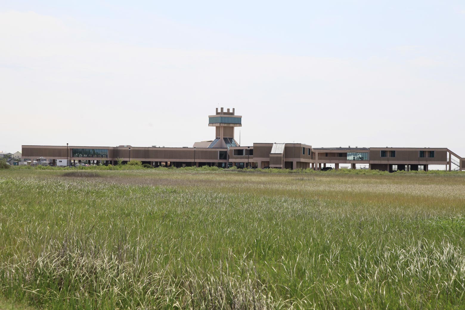 A field of grass in the foreground with a large brown building in the background. The building is one story, but raised on stilts. In the center is a lookout tower.