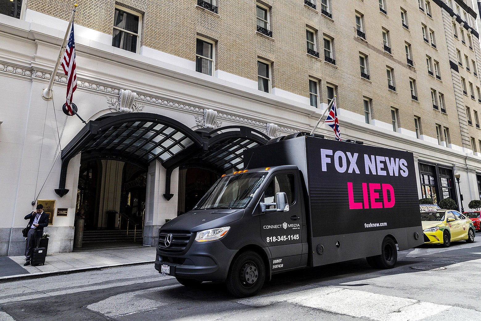 A black truck with a billboard on its side that reads "Fox News Lied." The truck is parked outside of a large hotel.  