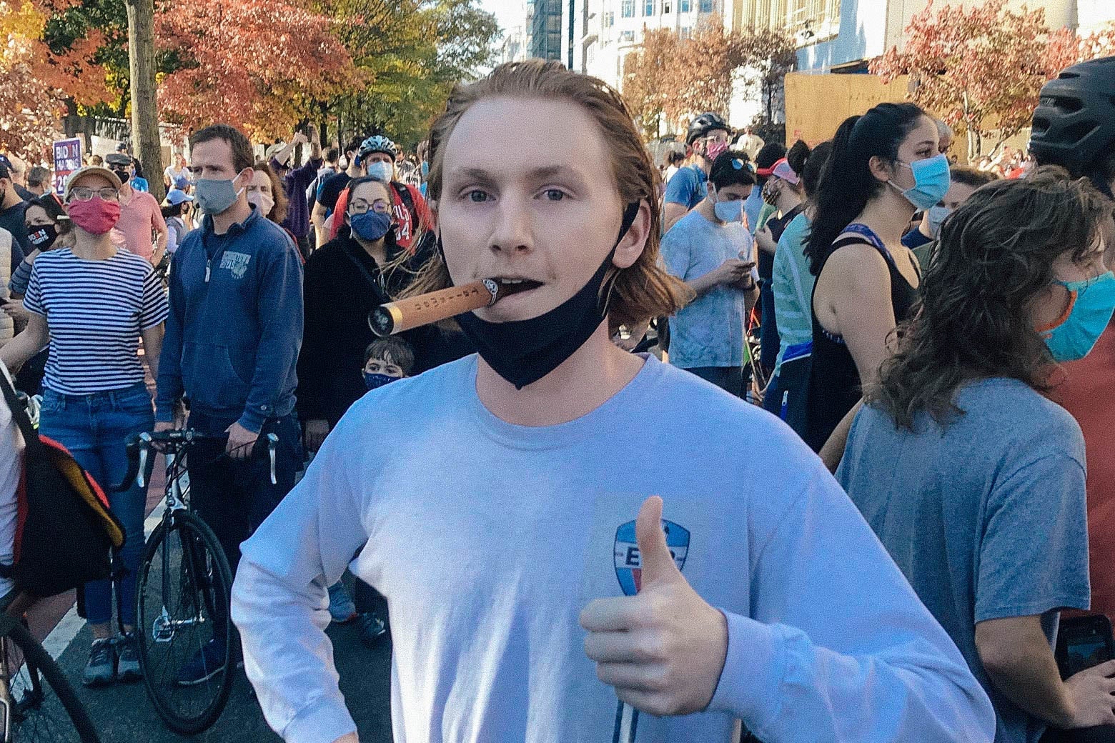 A man with a cigar dangling from his mouth gives a thumbs up, a black face mask around his chin.