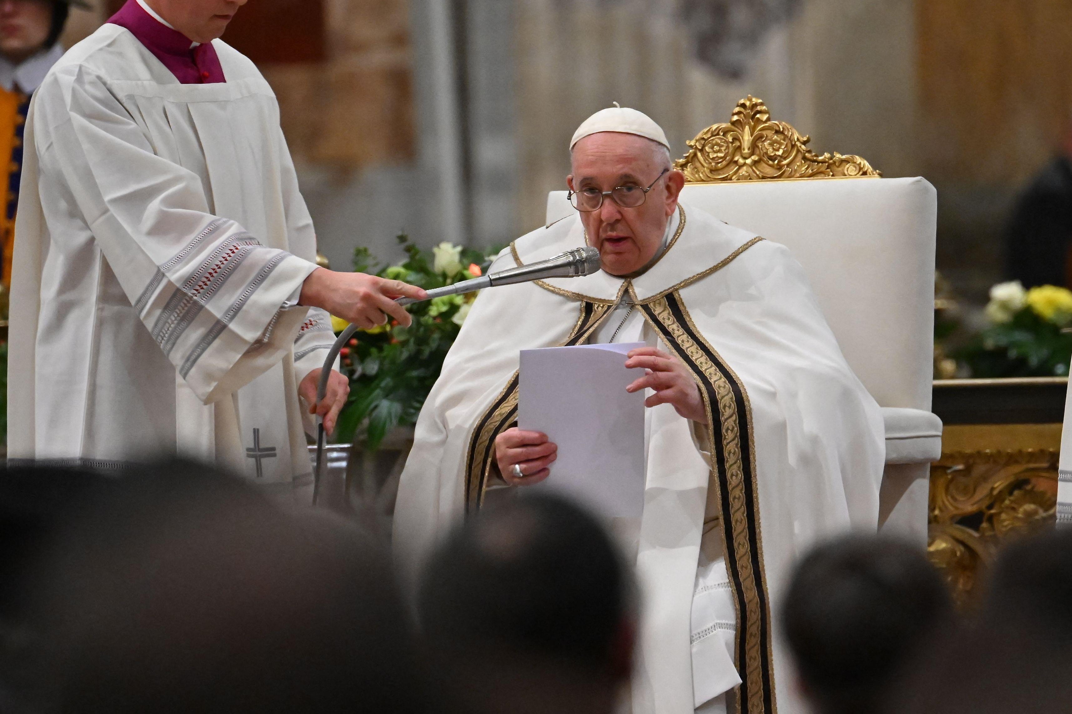 The pope, wearing white garments with black and gold trim and holding papers to read off, sits in a white chair with ornate gold trim and speaks into a microphone held by a man standing next to him. 