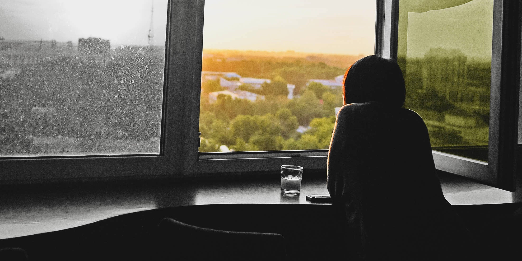 A woman staring wistfully out of a window as the sun pours through. A glass of water is on the sill in front of her.