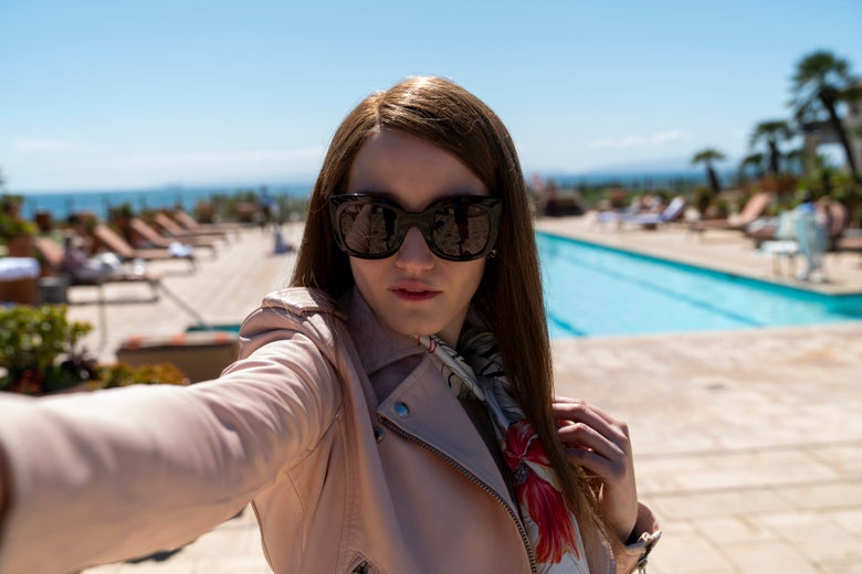 Julia Garner as Anna Delvey in a pink jacket and big sunglasses taking a selfie by a swimming pool