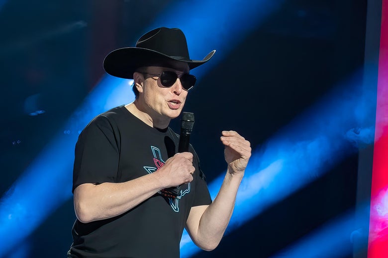 Elon Musk speaks onstage holding a mic in one hand while wearing sunglasses and a black cowboy hat