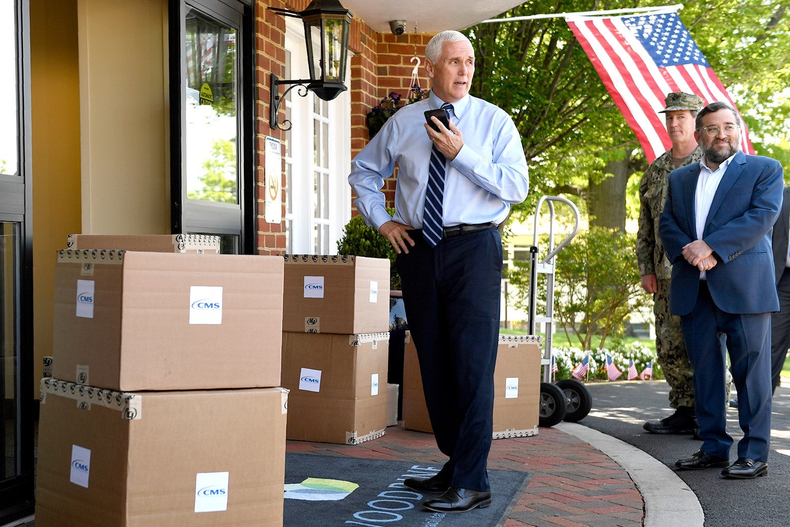 Pence, wearing a shirt and tie, speaks into a phone while standing next to piles of boxes.
