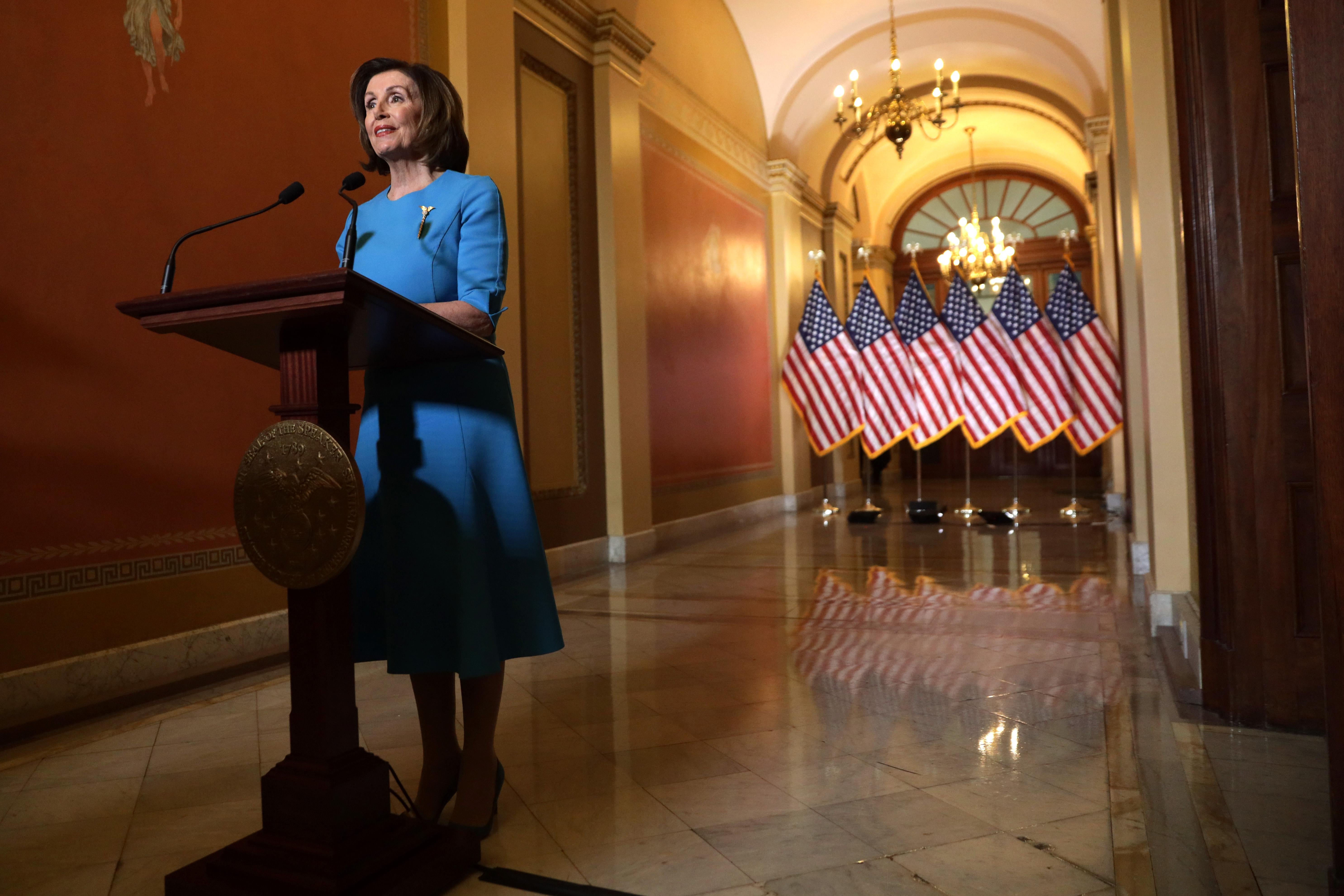 Nancy Pelsoi stands at a podium in a marble-floored hall with American flags in the background.