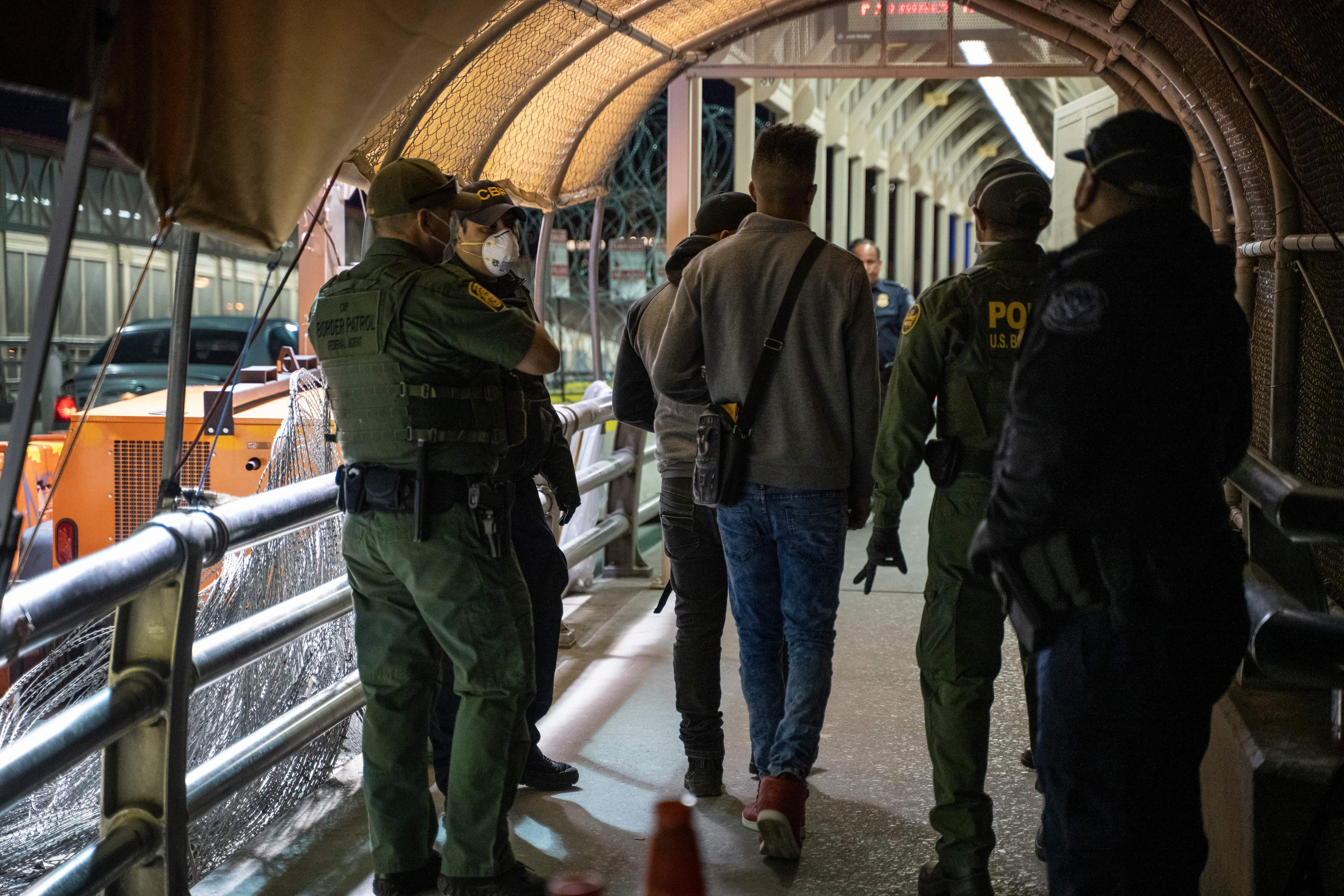 Uniformed CBP officers stand beside asylum-seekers in a covered walkway