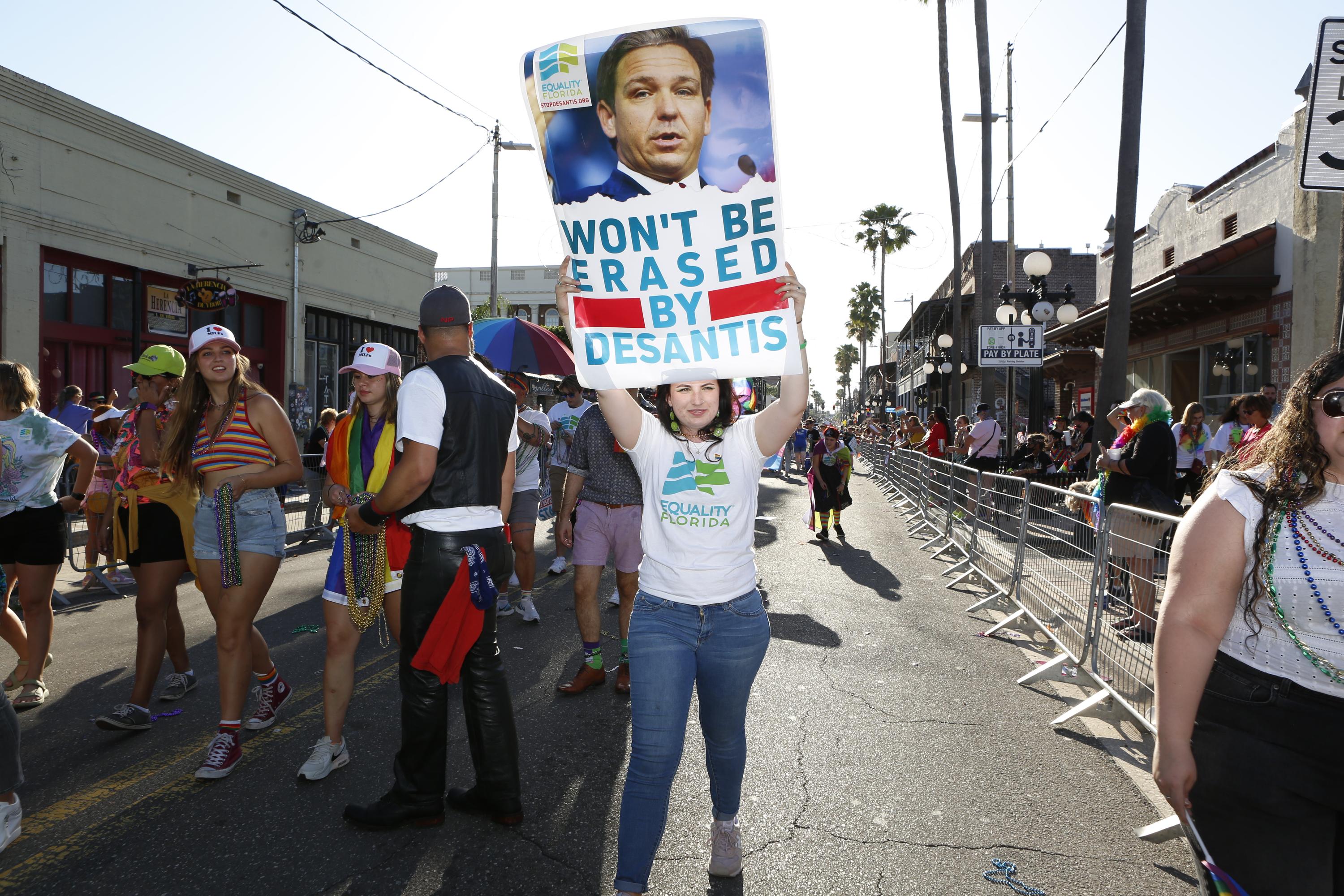 A person holds up a sign that says "Won't Be Erased by DeSantis" as they walk down the street with other parade participants.