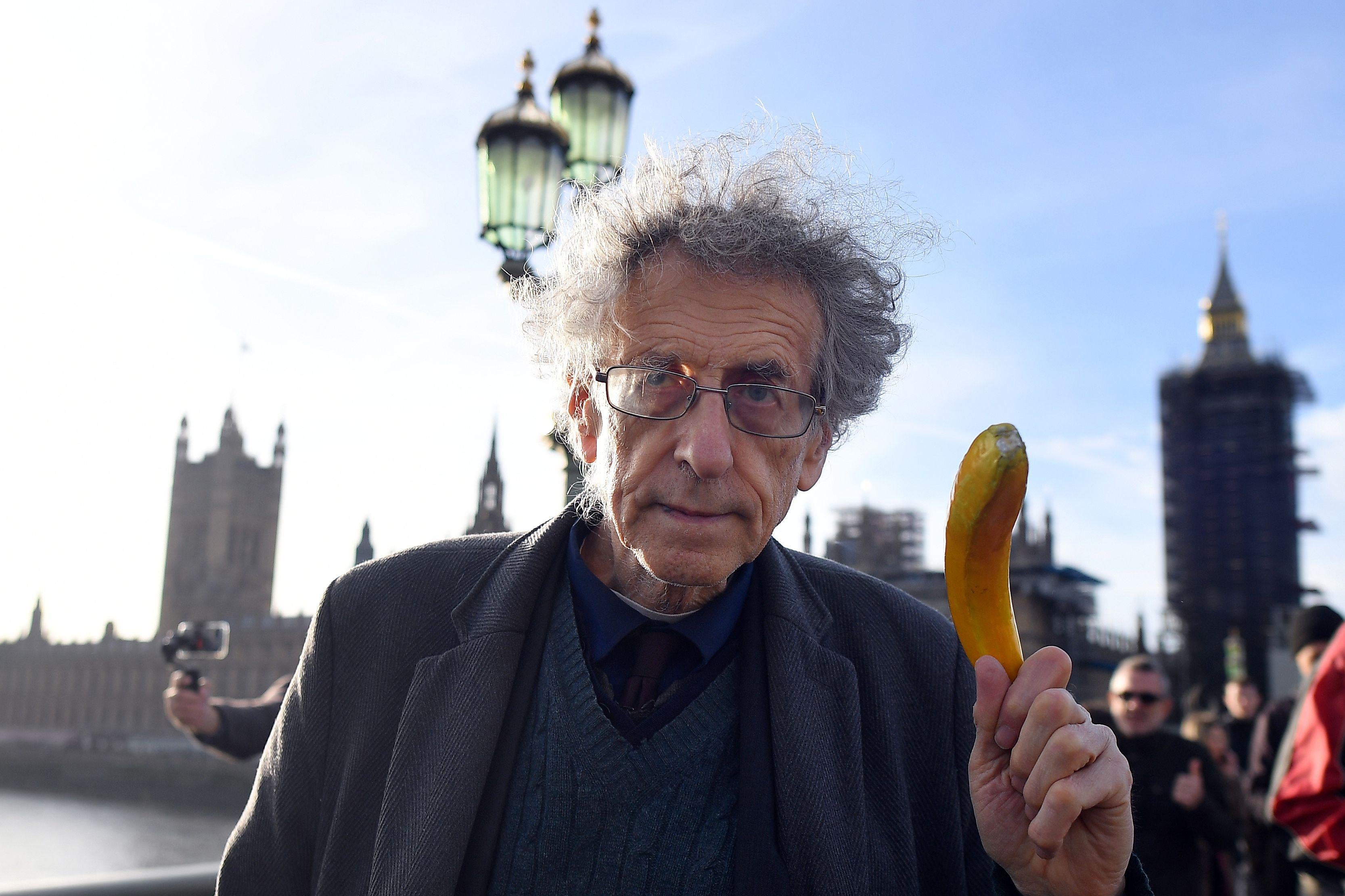 An older man with wild hair holds up a banana while standing outside in London.