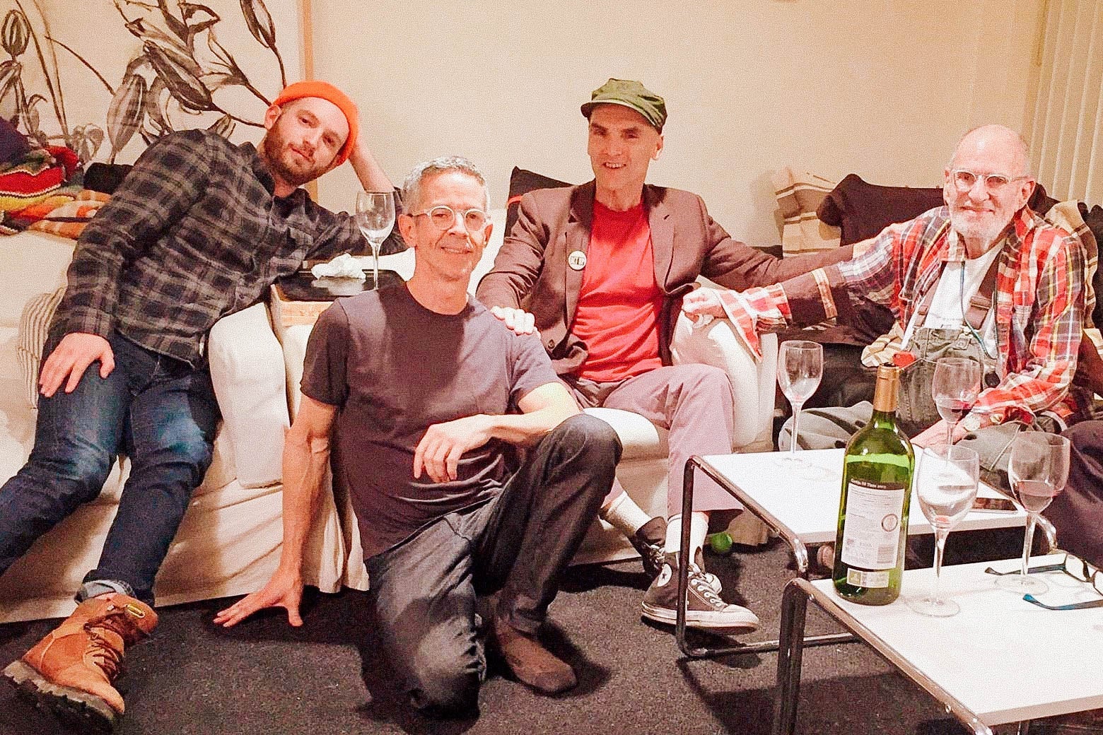 Four men pose together on sofas and chairs in a living room with a wine bottle and wine glasses on a nearby coffee table.