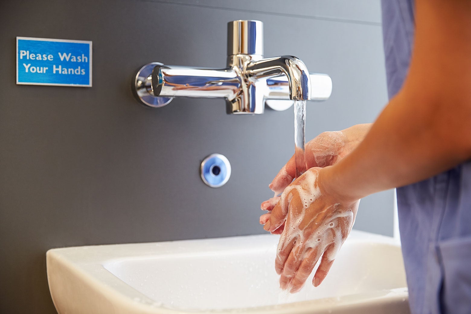 A nurse washing their hands in a sink next to a sign that says "Please wash your hands."