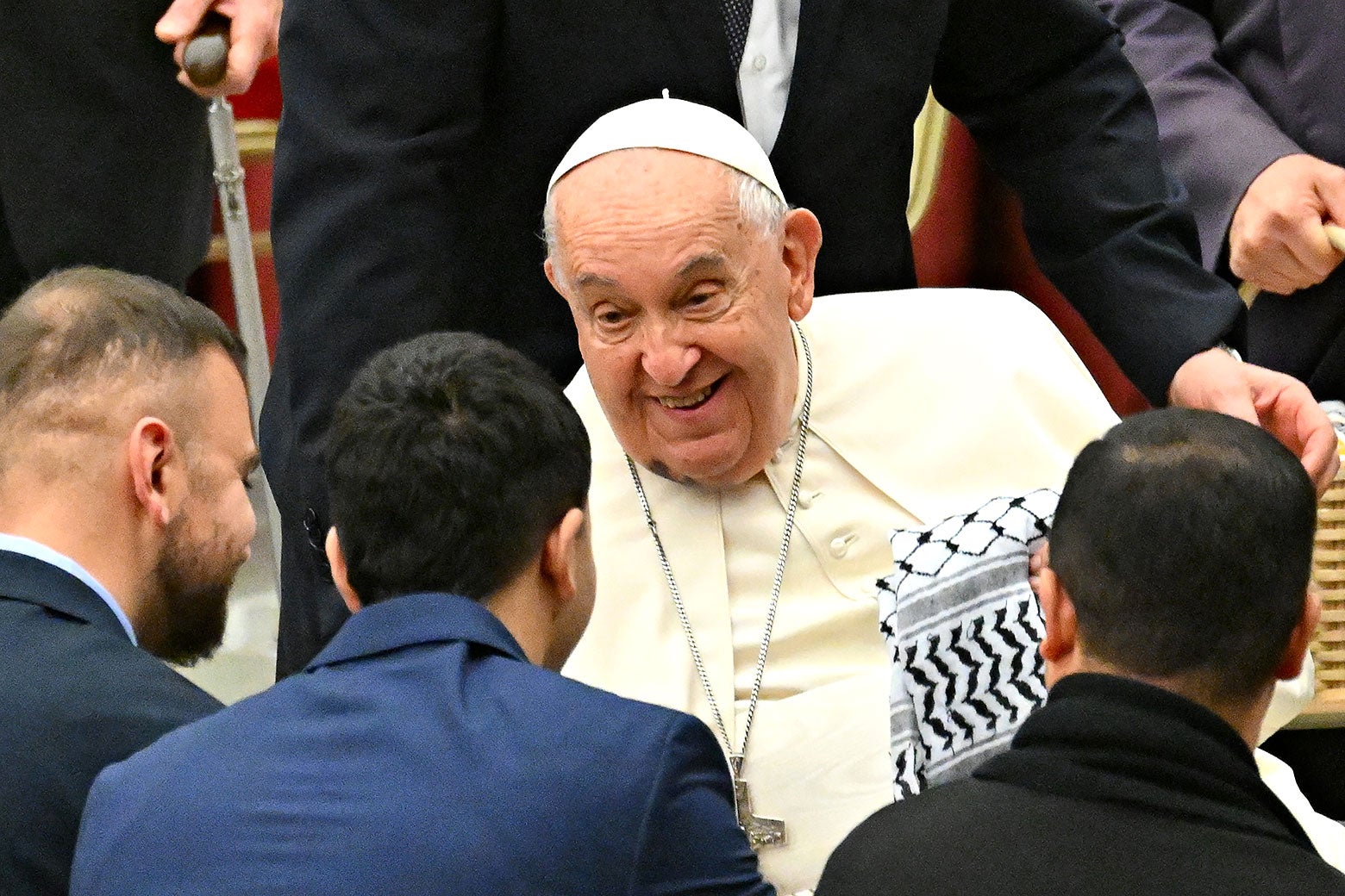 A man in white smiles and greets a group that hands him a kaffiyeh.