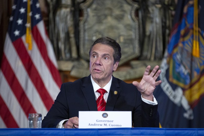 Cuomo, wearing a suit, gestures with his left hand while seated at a table in front of  U.S. and New York state flags during a daily coronavirus briefing in May.