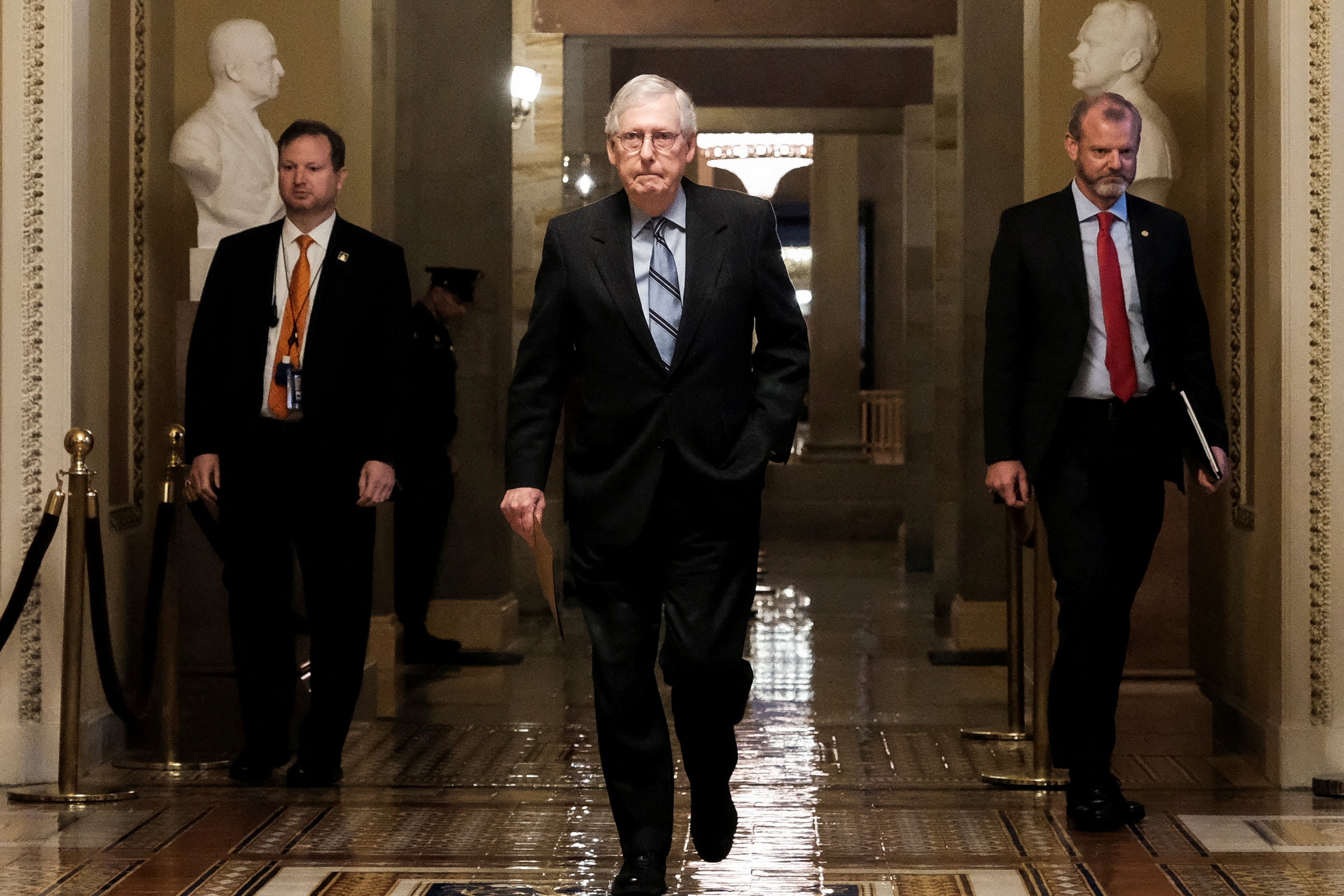 Mitch McConnell, flanked by two men, walks through the Capitol, with some statues behind him.