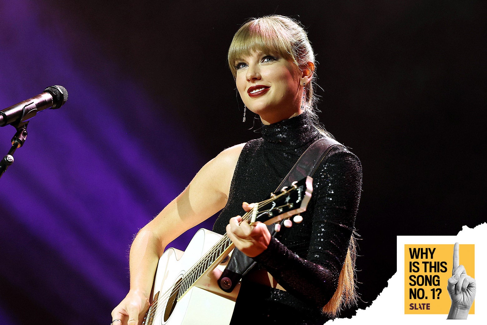 A photo shows Taylor Swift on stage smiling in a sparkly black shirt that has one long sleeve and otherwise shows one totally bare arm. In front of her, a white acoustic guitar, a microphone, and—photoshopped in the corner—a logo says "Why Is This Song No. 1?" with a little hand making the sign for the number 1