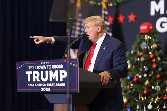 Trump shouting directly into a mic and pointing his arm straight ahead with a Christmas tree behind him.