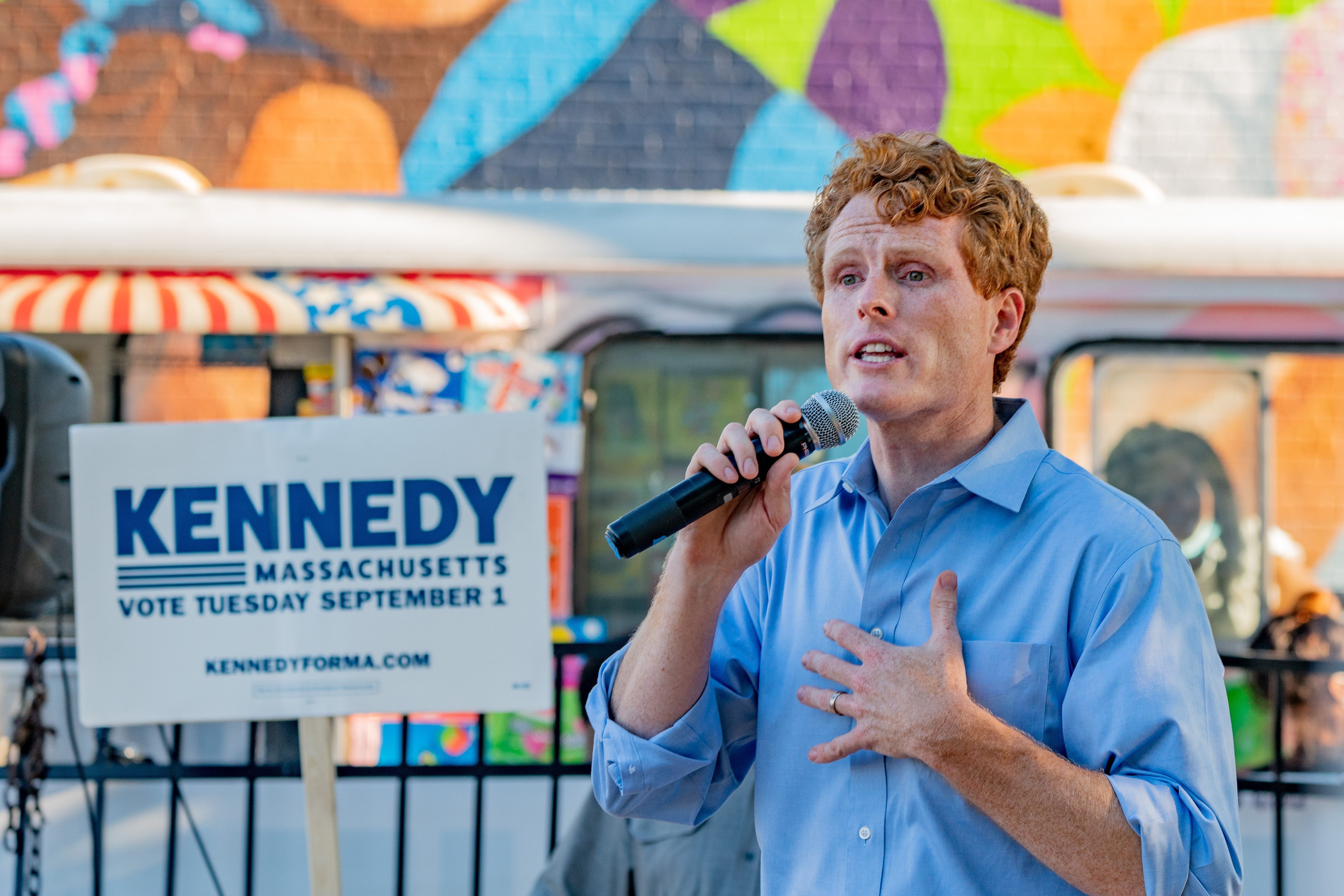 Joe Kennedy holds a microphone in one hand and his other hand to his chest while speaking in front of a mural.