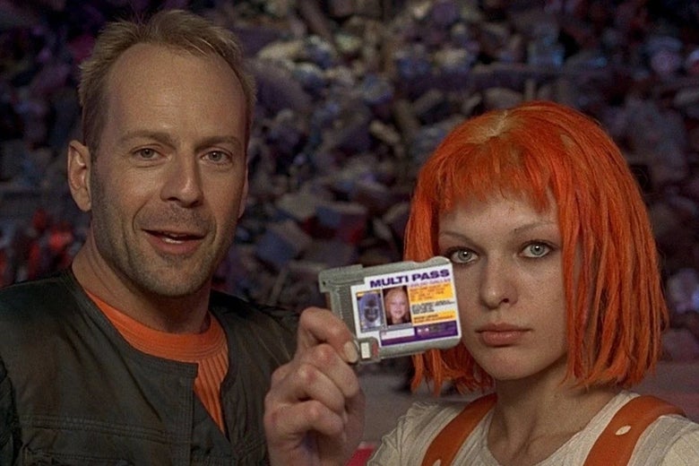 Bruce Willis stands smiling next to Milla Jovovich holding up an ID badge with fiery red-orange hair in a short bob-cut.