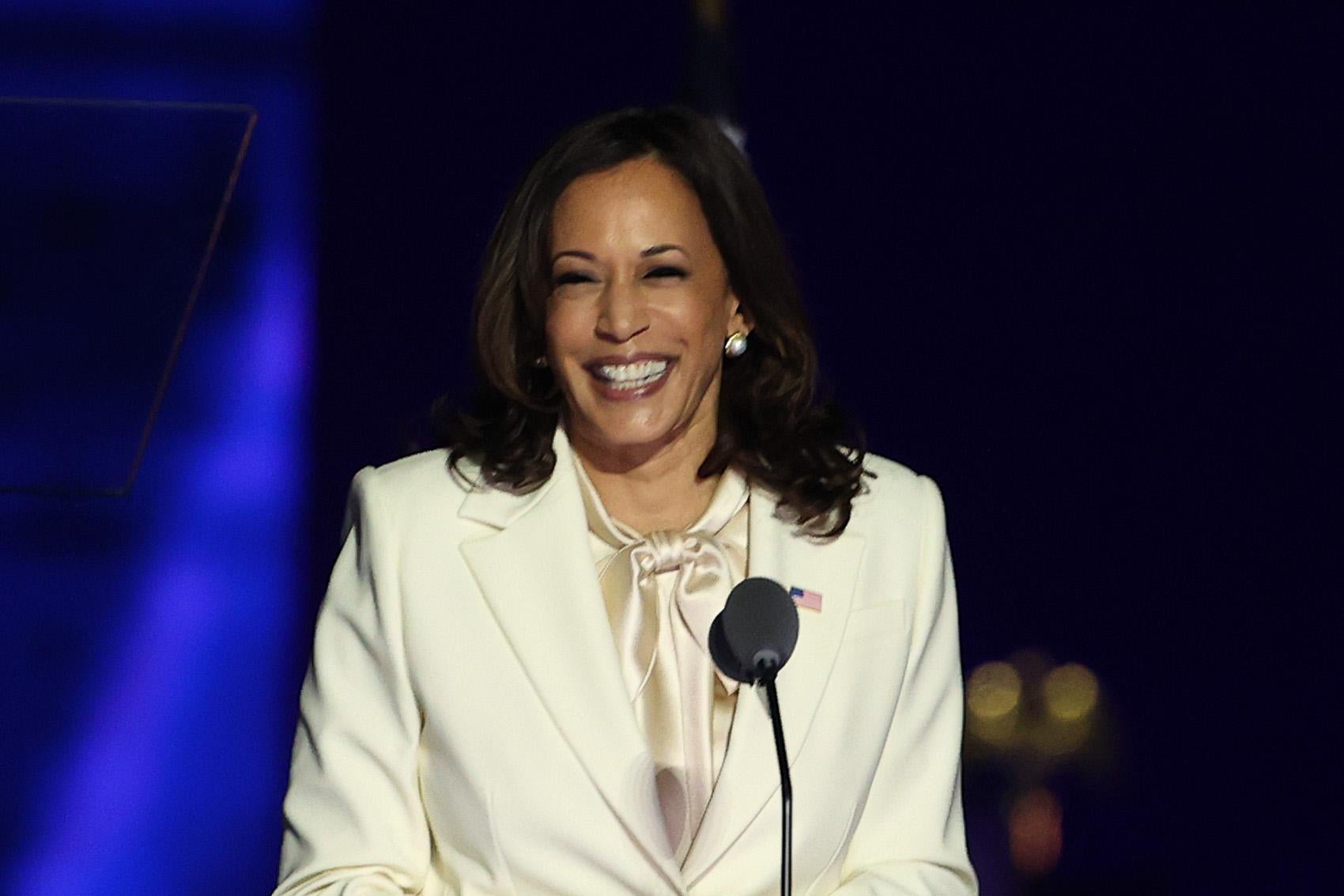 Kamala Harris, wearing a white pantsuit, grins onstage at a podium