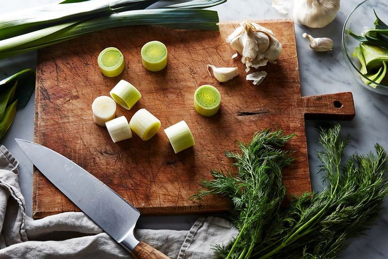 A cutting board laden with sliced leeks, garlic cloves and a garlic bulb. Whole leeks and another garlic bulb peek out from the top, fresh dill comes up from the bottom. A knife rests on the cutting board.