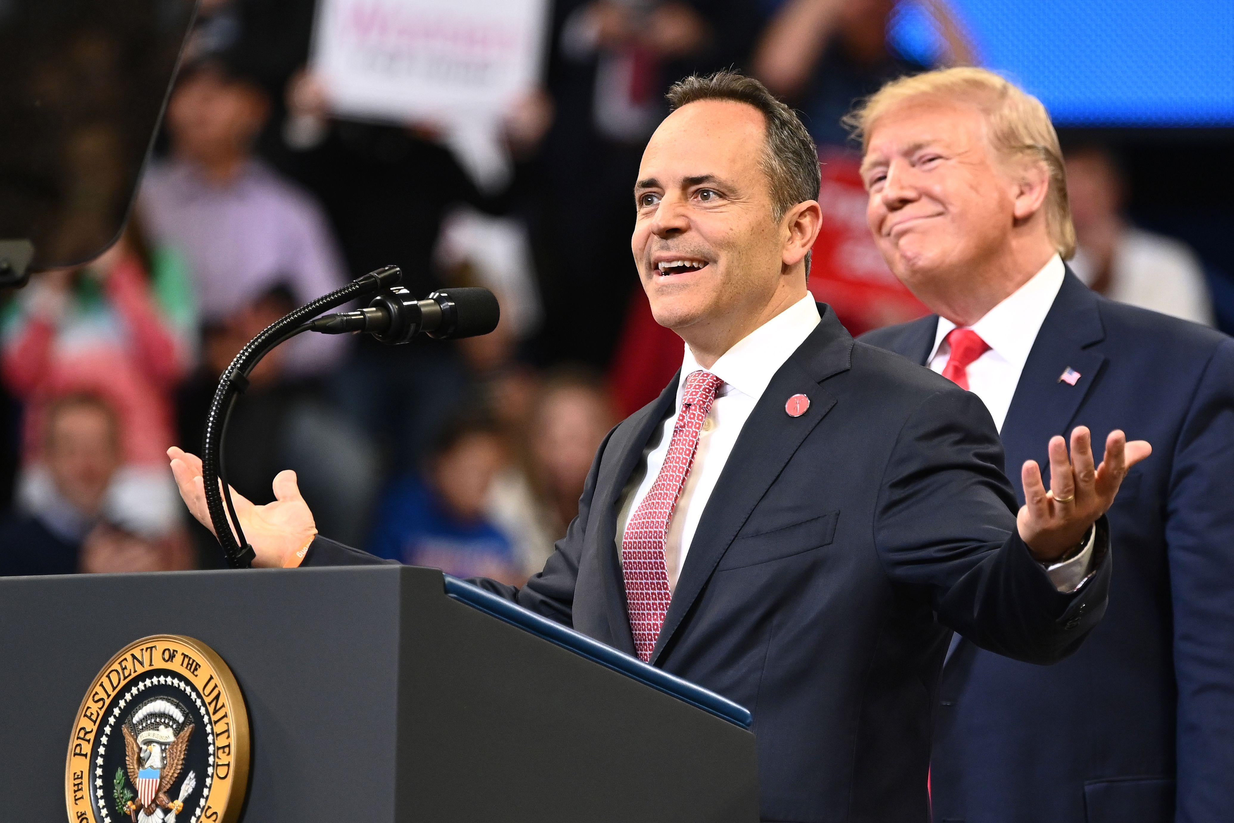 Trump smiles while standing behind Matt Bevin, who is giving a speech at a podium.