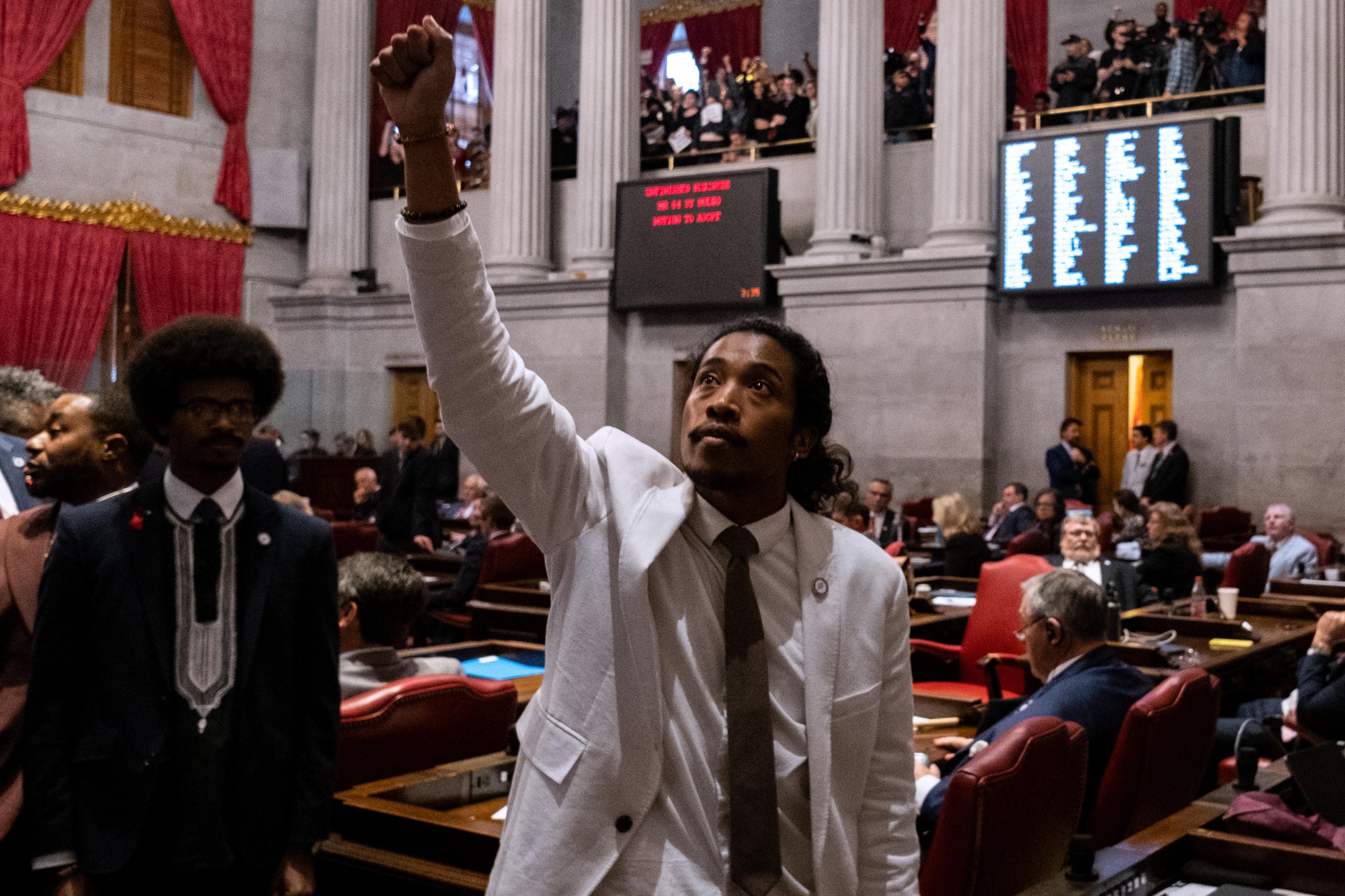 In the center of the frame, a Black man in a light suit, white shirt and brown tie stands with one fist raised on the State Capitol floor. To the left, a Black man in a blue suit looks on. Screens with the vote totals can be seen on the wall behind them, and other lawmakers are sitting and standing throughout the chamber in the background.
