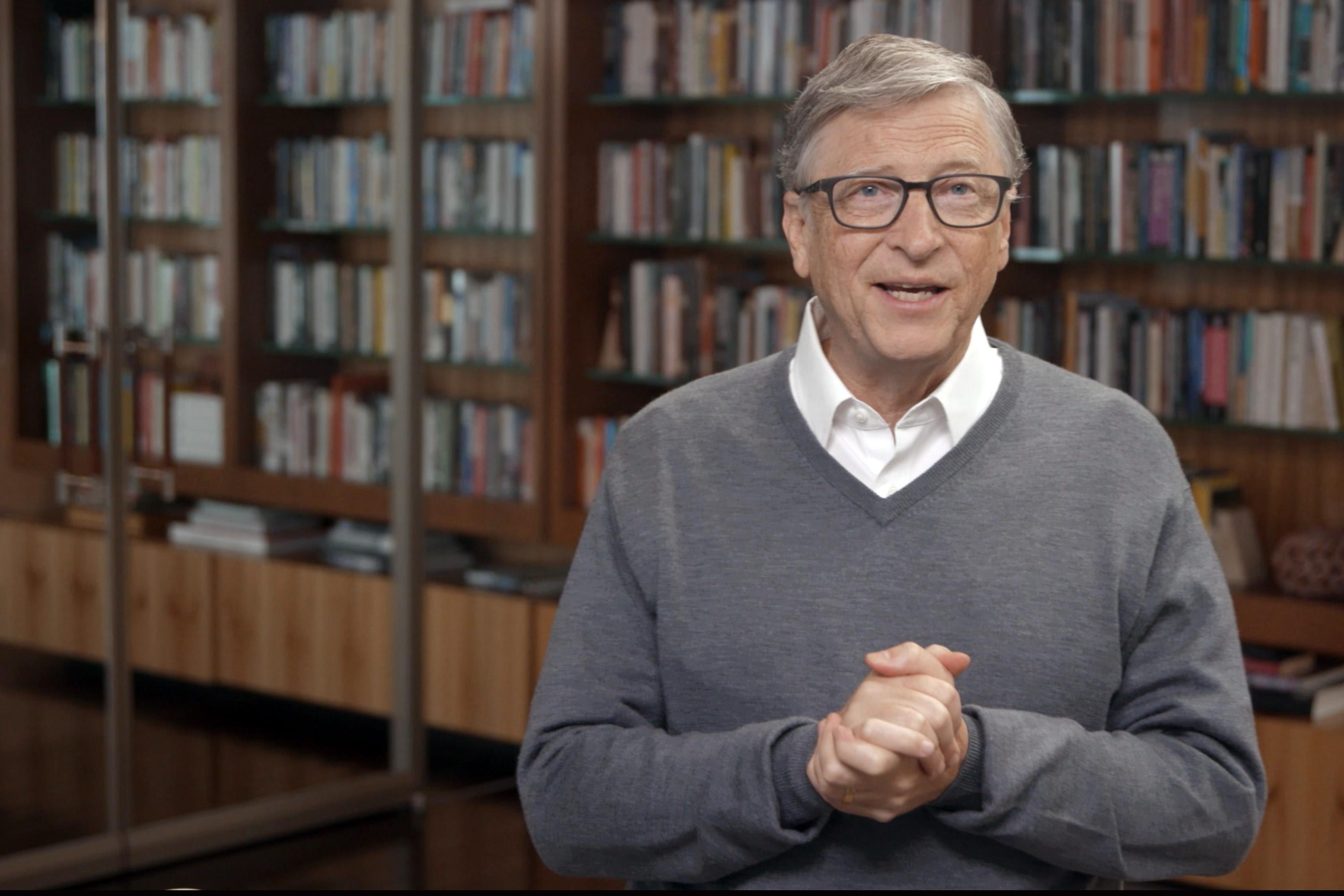 Bill Gates clasps his hands in front of bookshelves.