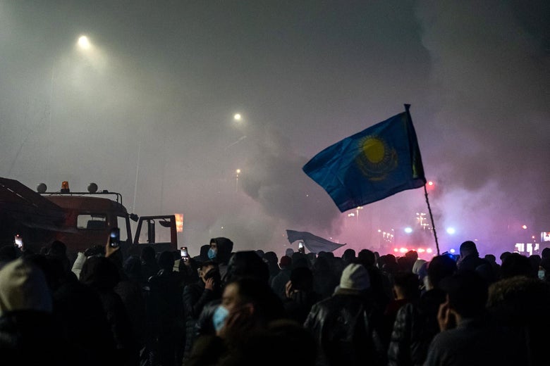 A Kazakh flag rises over a crowd during a smoky night.