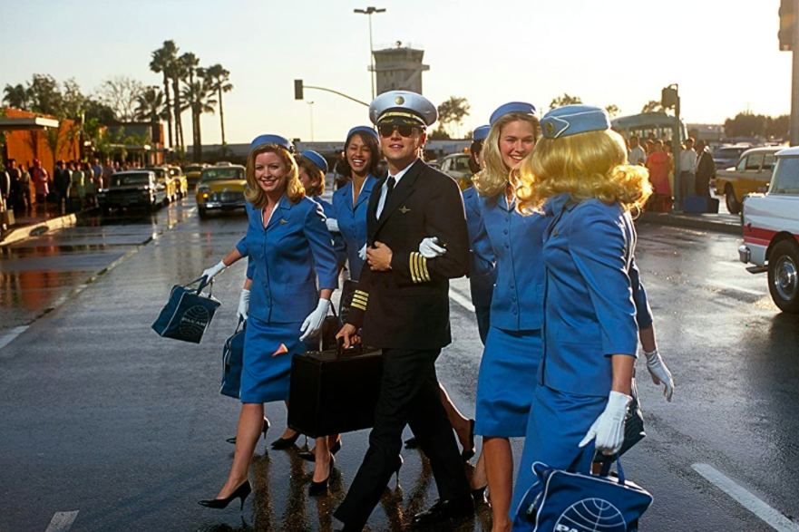 Leonardo DiCaprio in a pilot suit walks with a group of flight attendants.