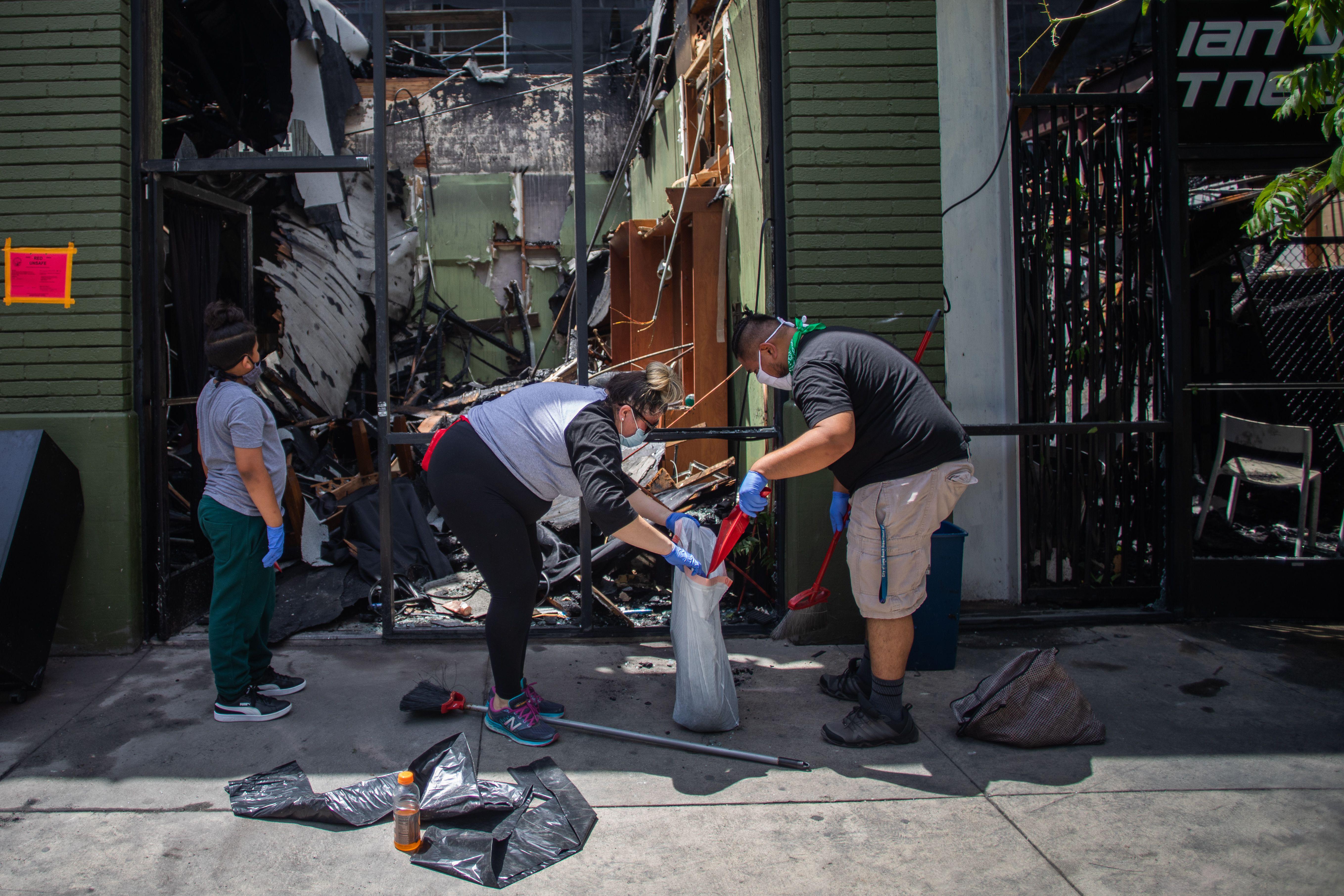 People wearing masks and gloves sweep debris into a garbage bag and survey the damage