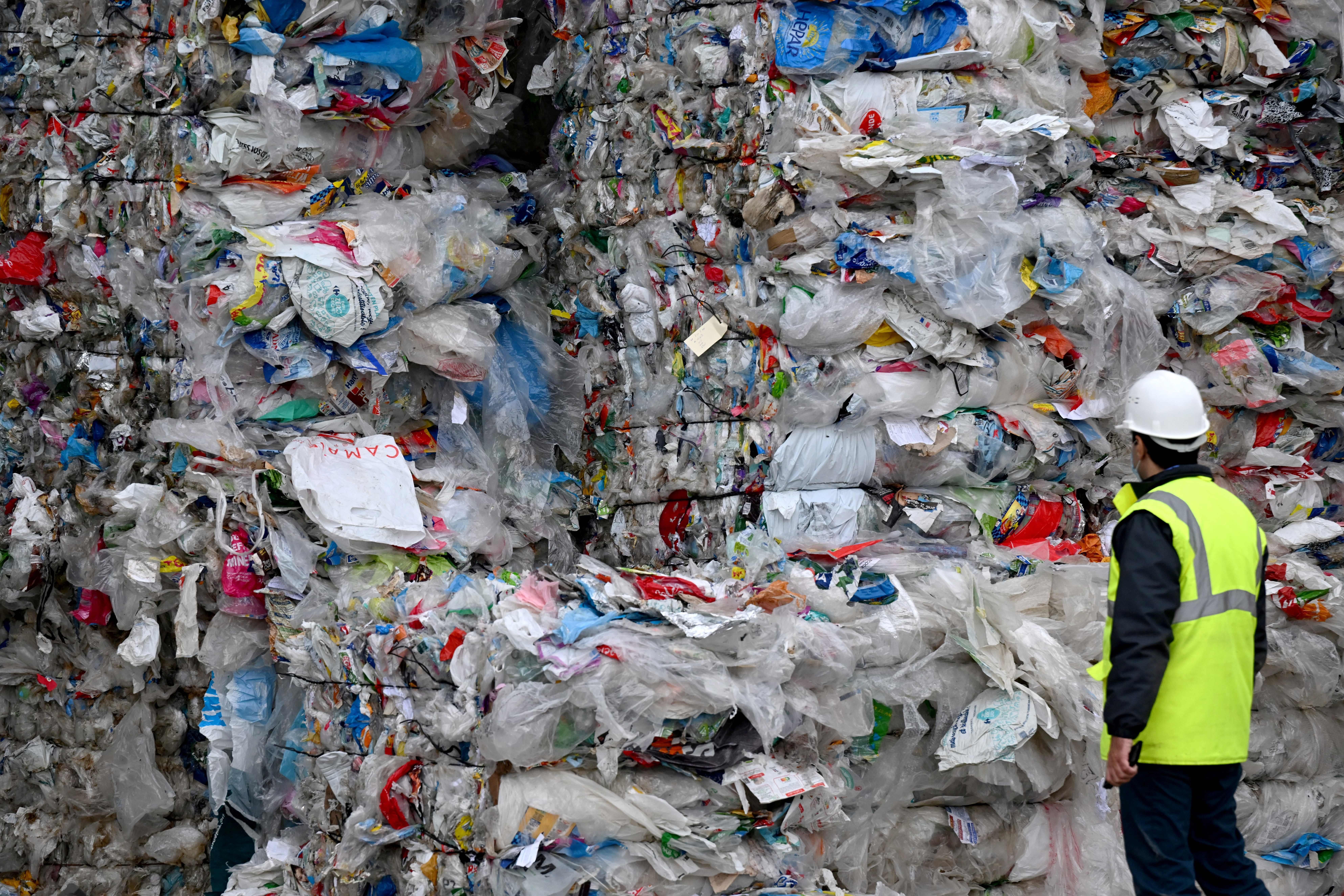 A man wearing a hard hat and yellow construction vest looks at towering stacks of bundled plastic waste.