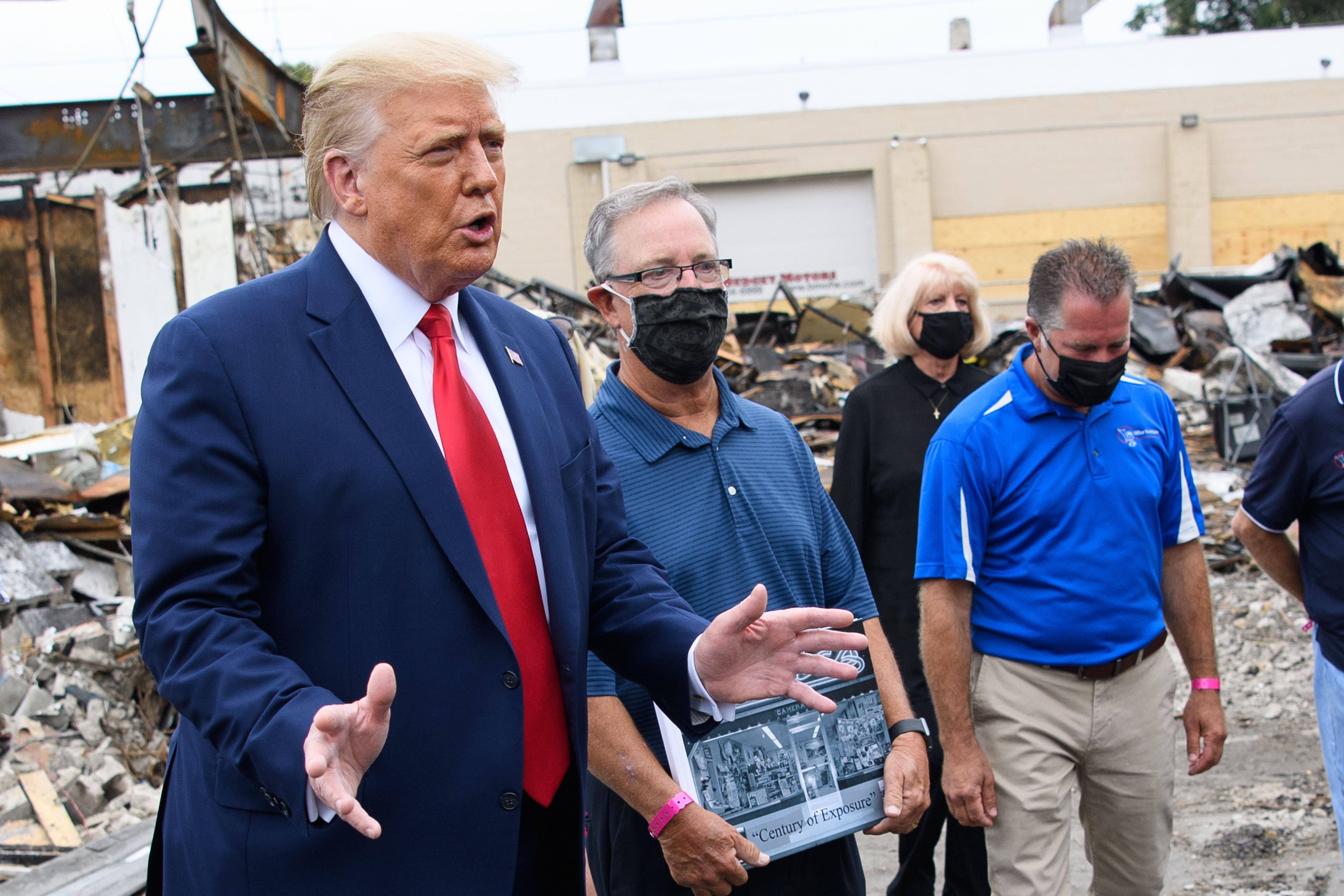 Trump gestures with his hands and speaks while standing amid rubble next to people in face masks.