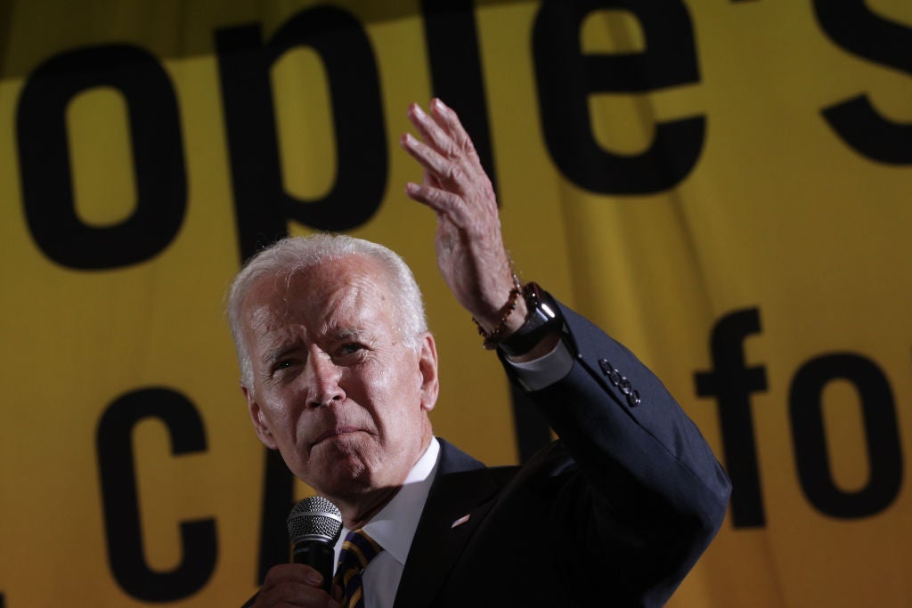 Joe Biden holds a microphone and gestures in front of a large banner.