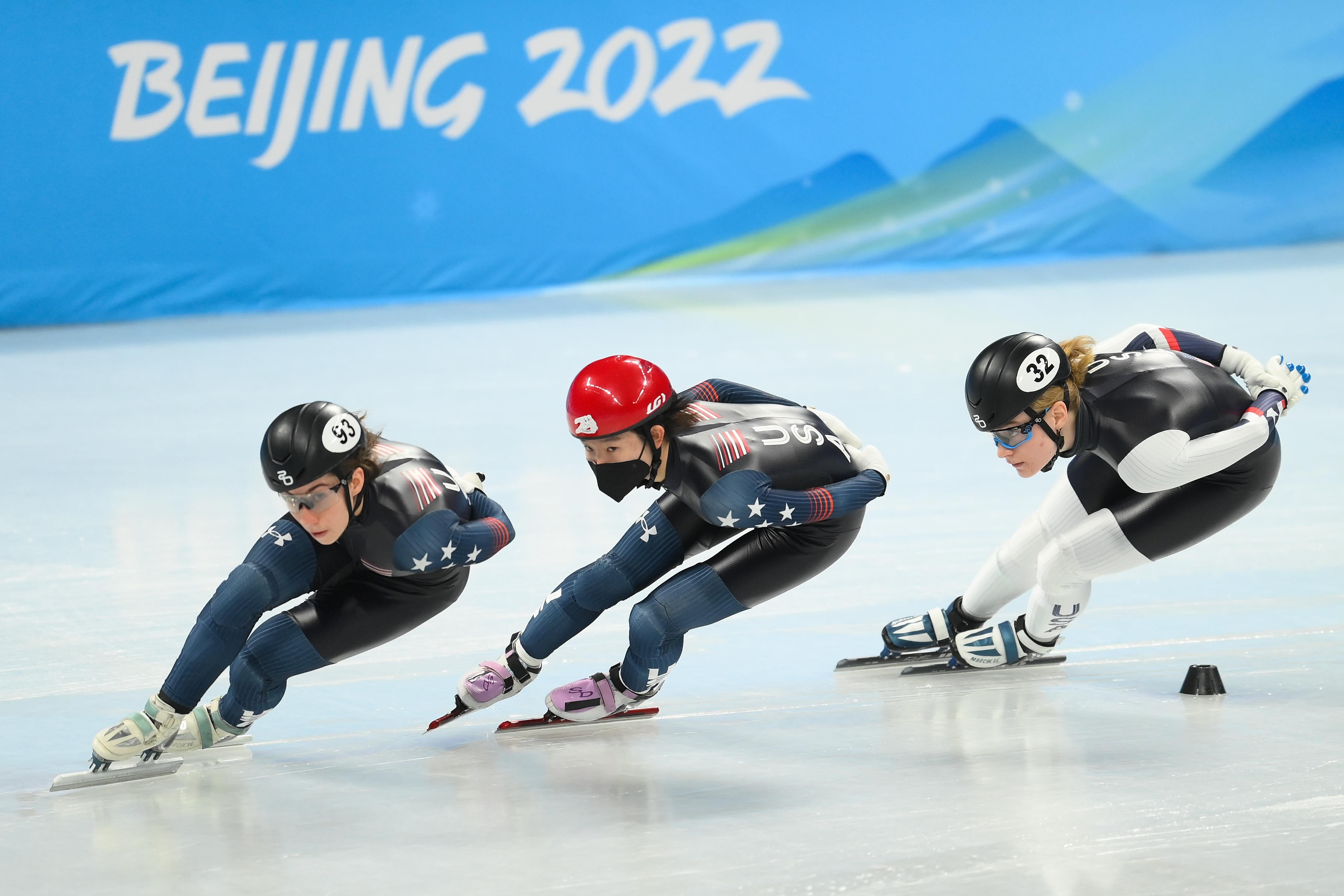 Three women speed skate in a single line with text on the wall behind them that says Beijing 2022.