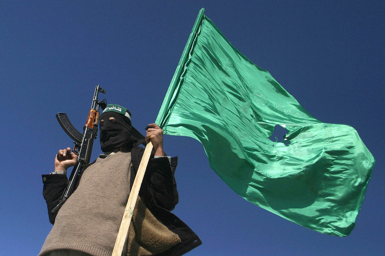A person with their face covered with a black cloth and wearing a green headband holds a gun in one hand and a green flag in the other.