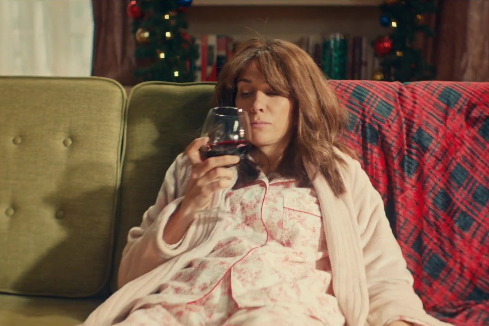 Kristen Wiig slouches on a couch in a living room covered with Christmas decorations, wearing a new robe, drinking a large glass of red wine.
