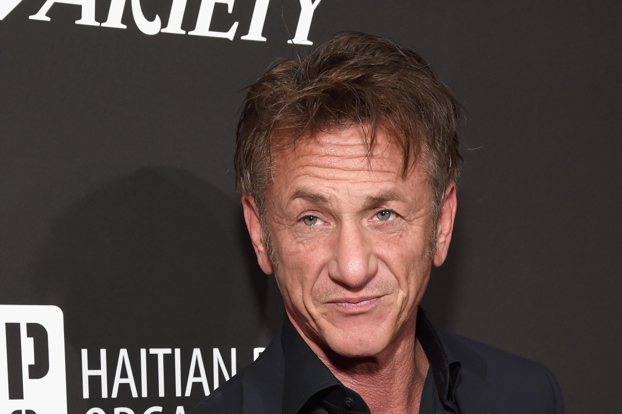 Sean Penn stands in front a black wall, wearing a black suit.
