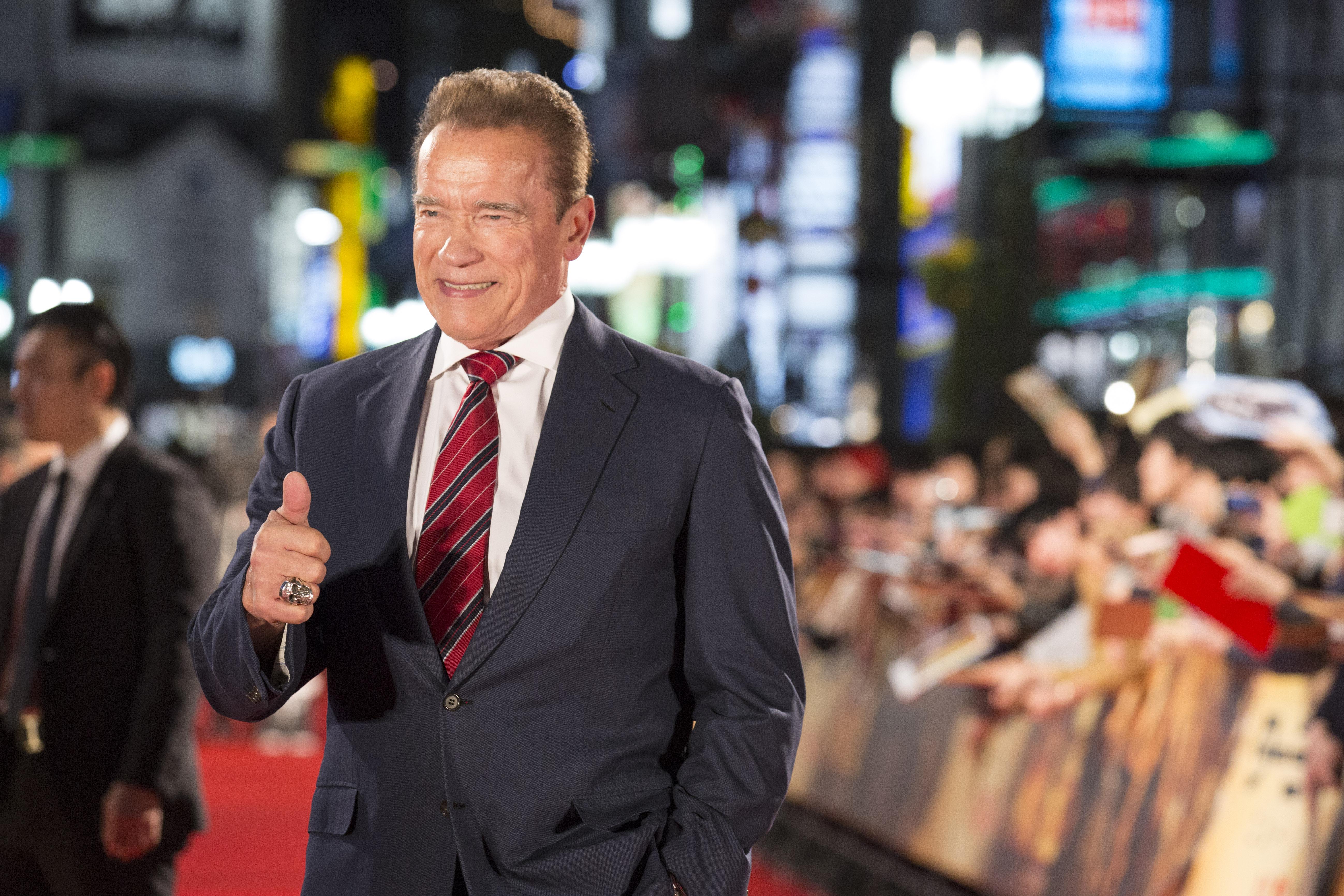 Arnold Schwarzenegger smiles and makes a thumbs-up on the red carpet, with cheering fans in the background