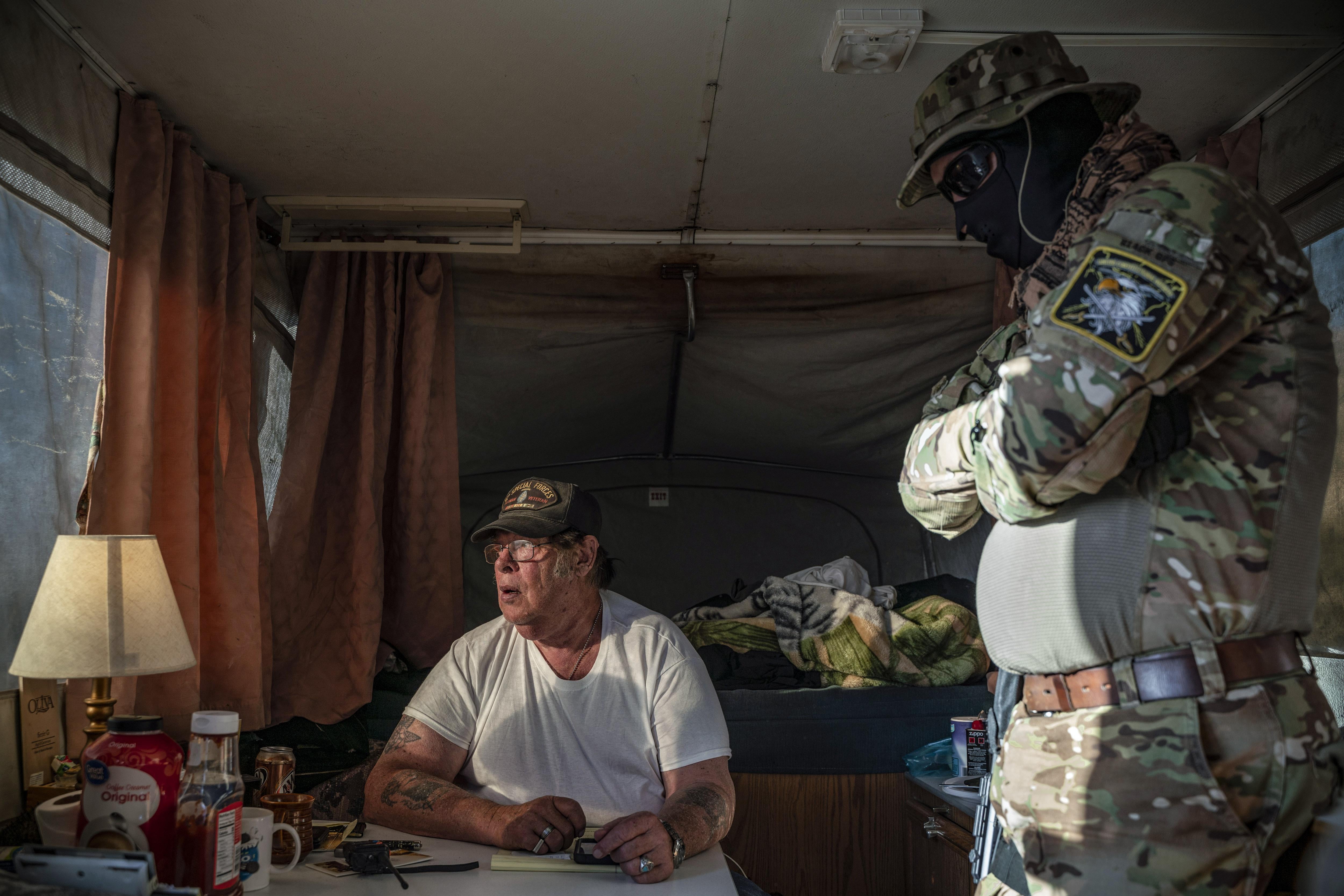 A man dressed in camo and a mask speaks with a man in a T-shirt inside a camper.