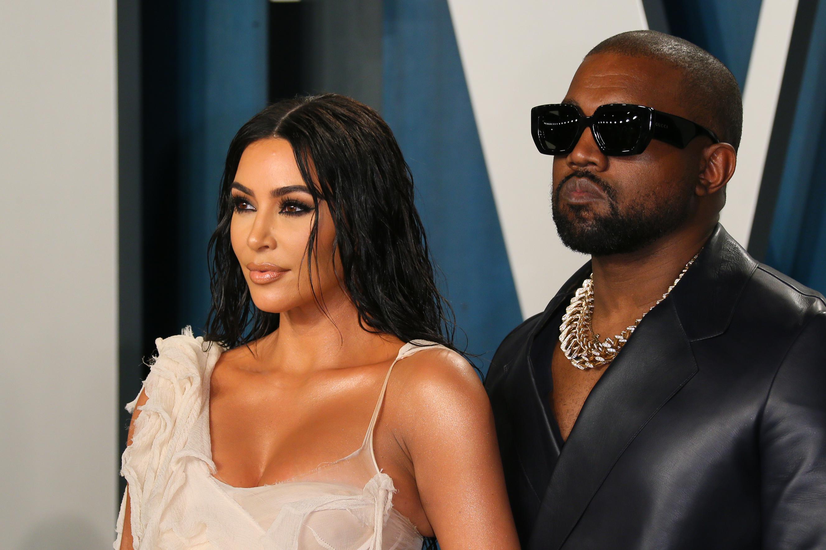 Kim Kardashian standing to the left of Kanye West, in formalwear in front of the Vanity Fair logo at the magazine's 2020 Oscar party. Both are seen from the chest up looking away from the camera, with hard to read expressions on their faces.