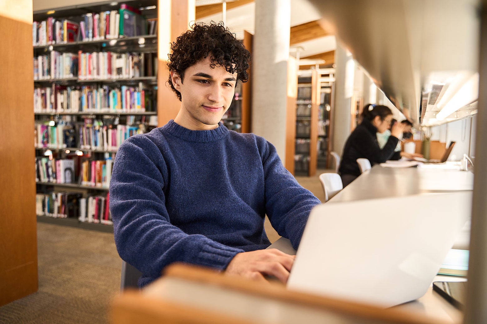 A college-age teen with dark curly hair sits in a library, typing on an open laptop in front of him.