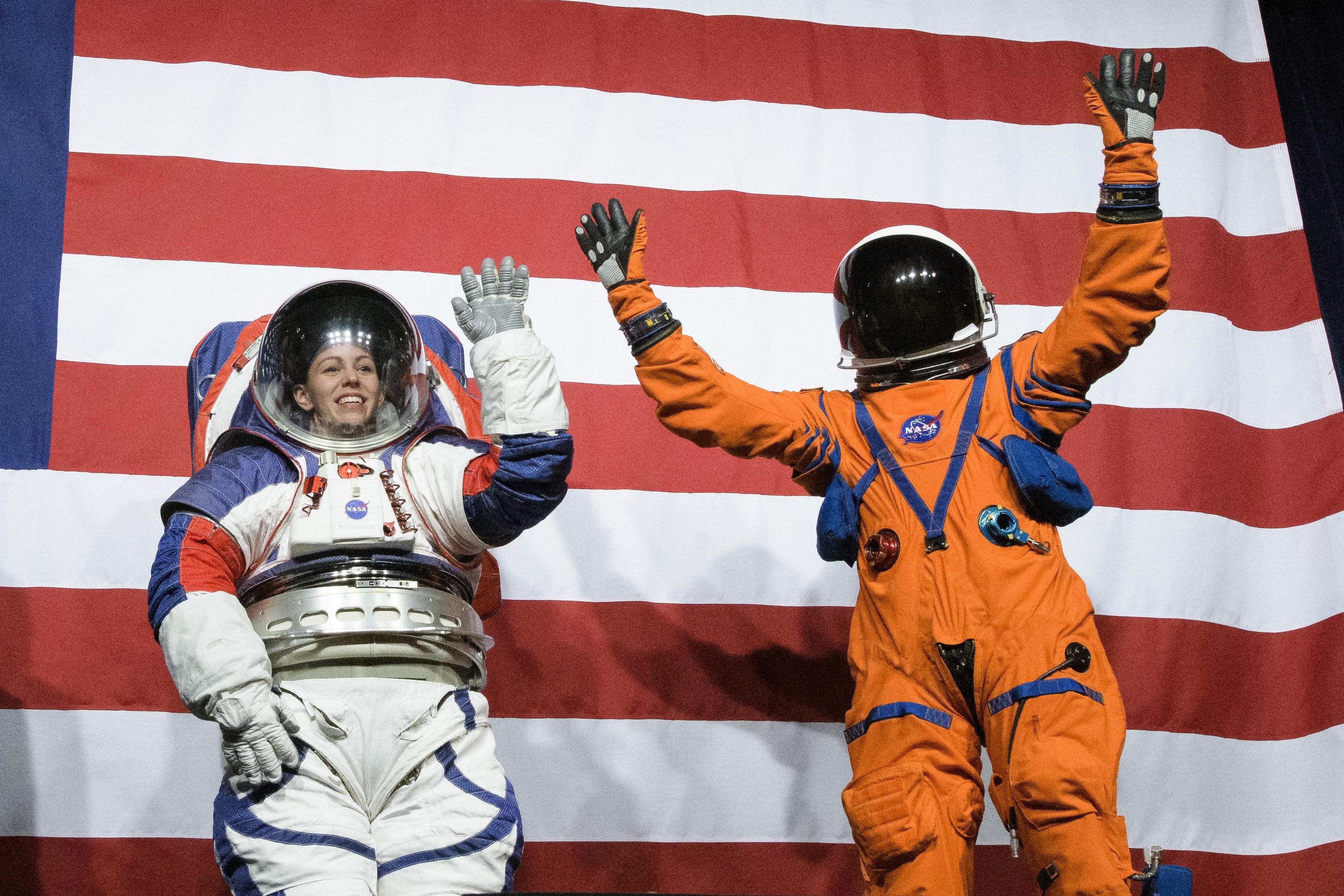 Two people stand in front of an American flag wearing spacesuits, with their hands raised in a waiving motion.